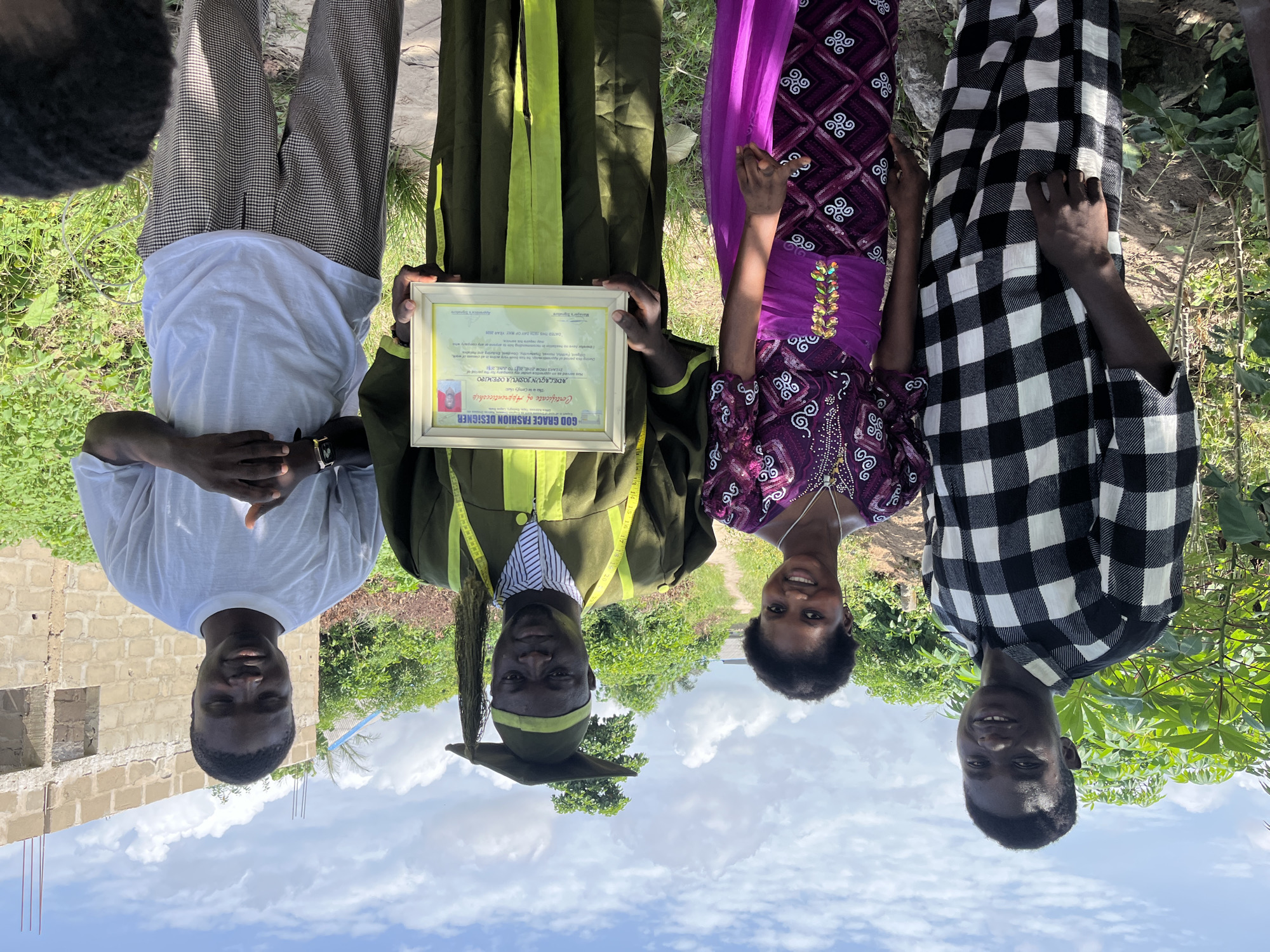 Four people are standing in a grassy area in Tofa, Nigeria. The person on the left is wearing a white shirt and checkered pants. The next person is wearing a green graduation gown and holding a framed certificate. The certificate reads "GOD GRACE FASHION DESIGNER." The third person is wearing a purple dress. The person on the right is wearing a black and white checkered shirt. The sky is blue with white clouds. A building is under construction in the background.