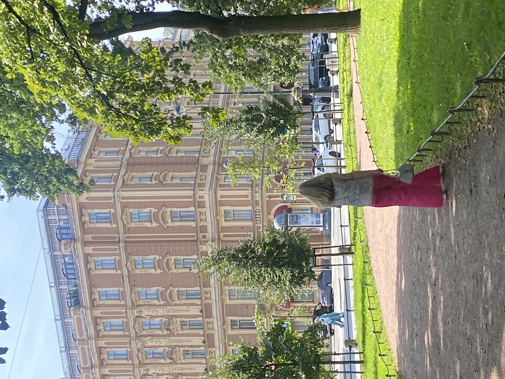 The image shows a woman walking on a path in a park in Saint Petersburg, Russia. She is wearing a grey patterned top and bright pink pants, carrying a light grey purse. In the background, there is a large, ornate, multi-story building with a tan facade and yellow trim. The building has many windows and decorative architectural elements. There is a street in front of the building with several cars driving on it. Green trees line the street and the park. The sky is blue with some wispy clouds. The overall scene depicts a sunny day in a European city.
