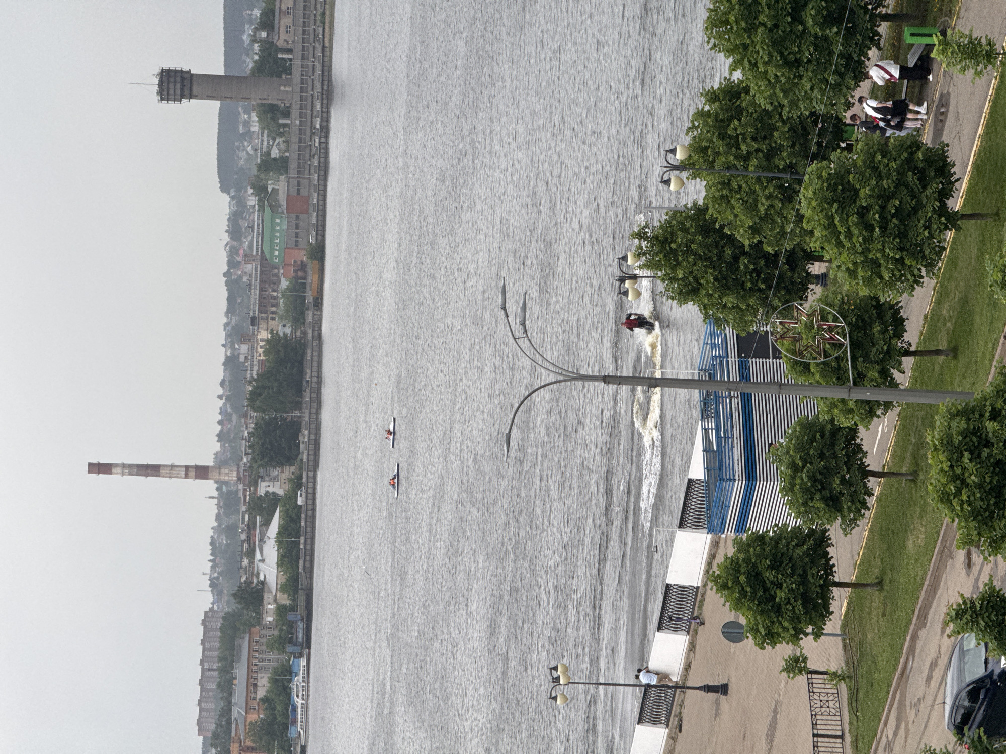 The image depicts a waterway in Izhevsk, Russia, with a jet ski, two canoes, and urban landscape elements.
The waterway is grey, indicating overcast weather, and shows a single jet ski leaving a noticeable wake, oriented toward the shore. Two canoes, each containing one person, are visible further out in the water.
The shore includes a walkway, trees, and streetlights. A small group of people are near the walkway.
The city backdrop includes a large chimney and a tall tower structure. The overall scene suggests a waterfront area within an urban setting.