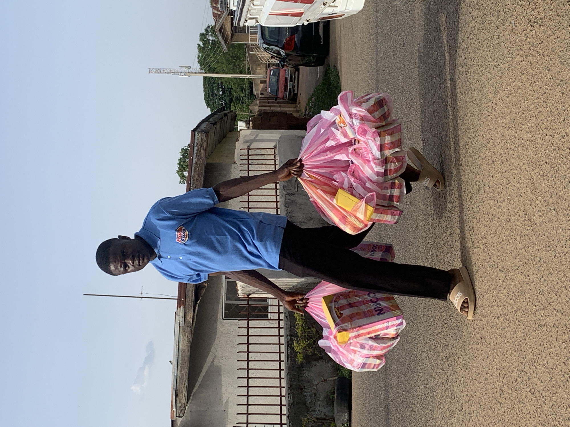 A man stands on a paved road in Jos, Nigeria. He wears a short-sleeved blue shirt with an "HANS & SONS" logo, black pants, and beige Adidas slides. He carries two large pink and white striped plastic bags filled with boxes of corn snacks in each hand. The background includes buildings, a utility pole, parked cars, and green trees. The sky is light blue.
