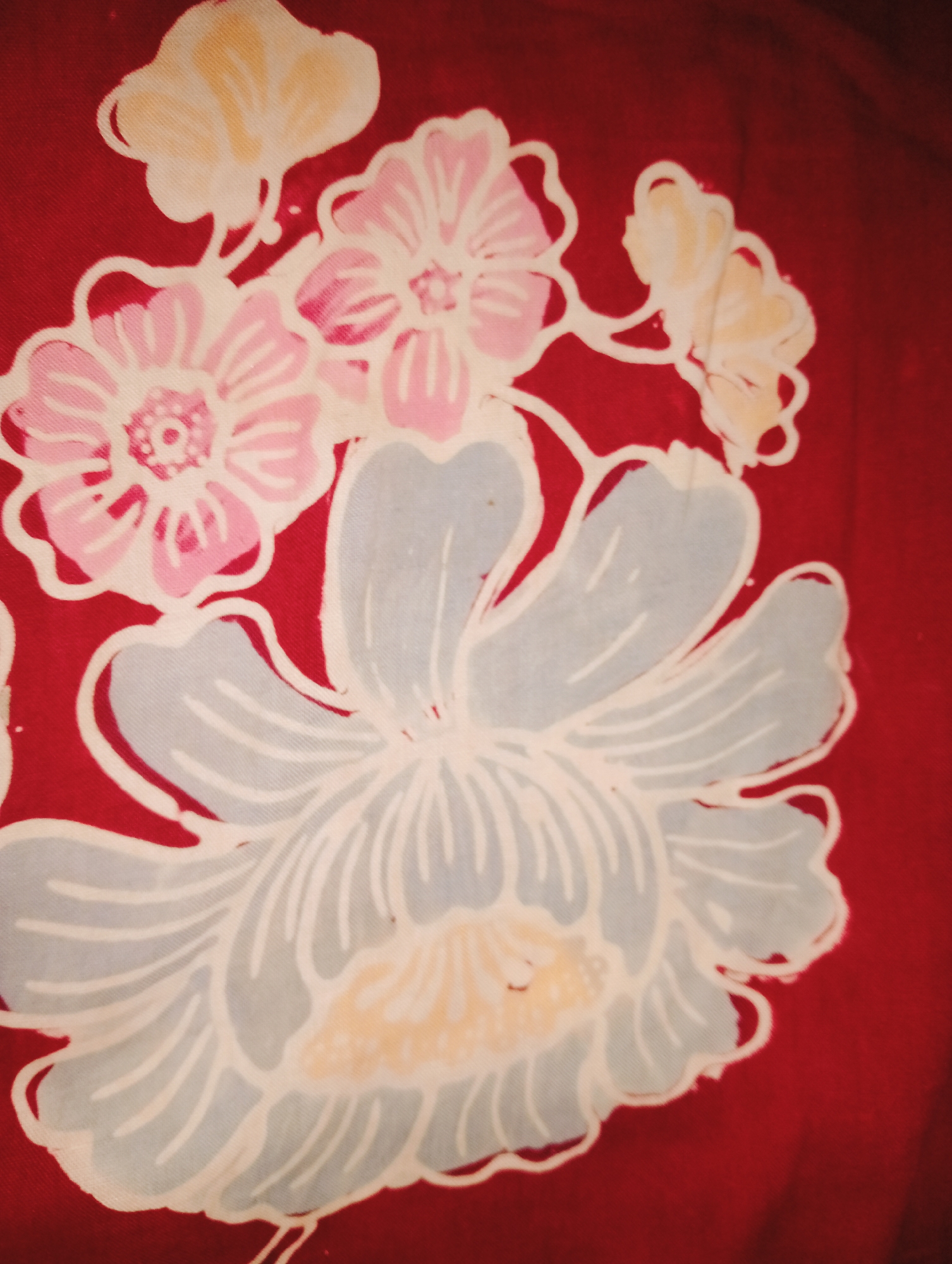A close-up photograph shows a section of red fabric with a floral pattern. The flowers are rendered in a stylized manner with outlines in white and fill colors of light pink, light yellow, and light blue. The largest flower, in the foreground, is light blue with white petal outlines and a light yellow center. Behind it, a pink flower with white outlines is visible, and further back are smaller, lighter yellow floral elements. The fabric appears to be silk or a similar material, with a slight sheen. The image is cropped and does not provide any contextual information about its location in Tridayasakti, Indonesia.