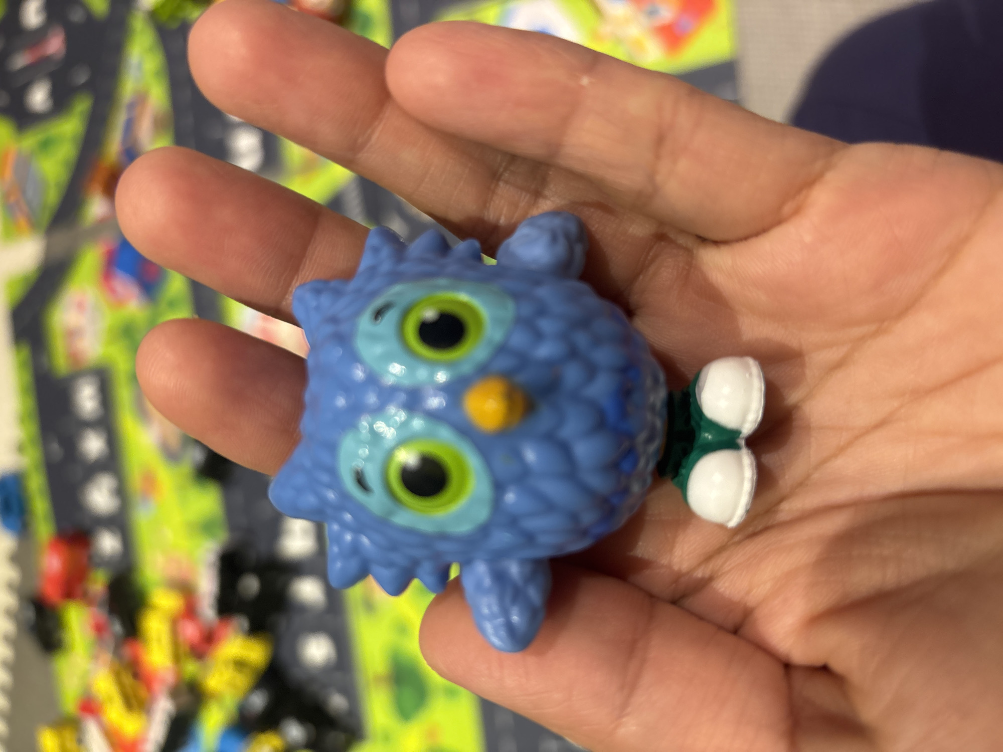 A close-up, eye-level shot shows a person's hand holding a small, blue, cartoonish owl figurine. The figurine has large, green eyes with black pupils, a small orange beak, and is wearing green shorts and white shoes. The owl's body is textured to resemble feathers. The background is out of focus and appears to be a colorful playmat with images of roads and toy vehicles, suggesting a child's play area. The lighting is bright and even, with no distinct shadows. The image is crisp and sharp, with the figurine being the primary subject. There is no discernible location in Dubai, United Arab Emirates.