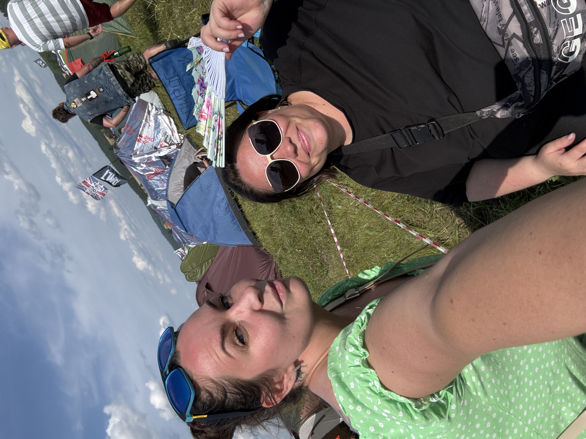 Two women are in a grassy field with tents and flags in the background, suggesting a festival or outdoor event. The woman in the foreground on the left is wearing a green dress with white polka dots and blue sunglasses. She is taking a selfie, with her arm extended. The woman on the right is wearing a black t-shirt and large cream-colored sunglasses, holding a fan. The environment appears to be a campsite under a partly cloudy sky. The image was taken in Lagunovo, Russia.