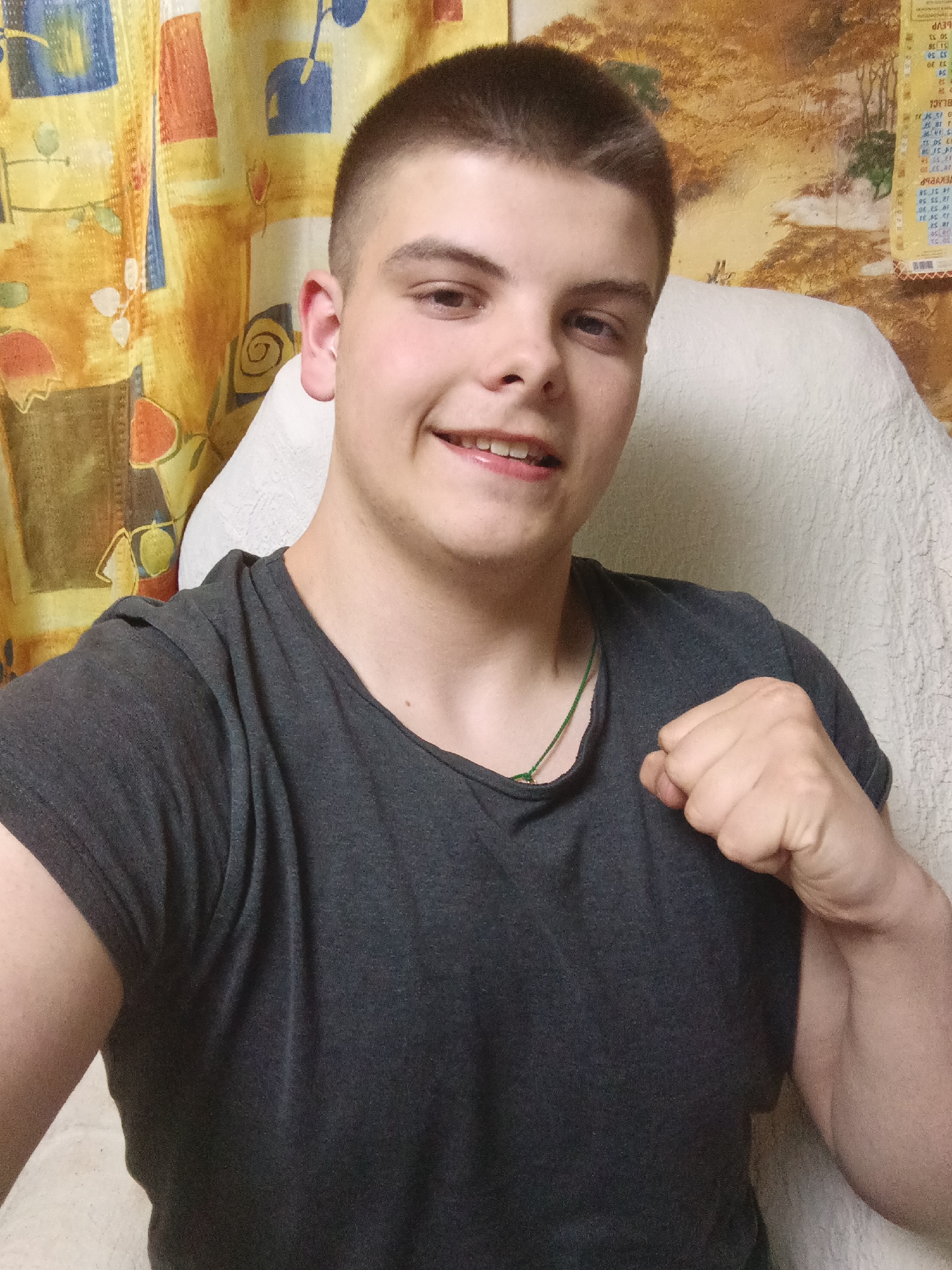 A young man with a buzz cut is posing for a selfie. He is wearing a dark grey t-shirt and a green necklace. He has a slight smile on his face and his right hand is clenched in a fist near his shoulder. The background includes a yellow patterned curtain and a white, textured chair. A calendar with Chinese writing is visible on the wall. The location is likely Красный Маяк, Russia, based on the request.