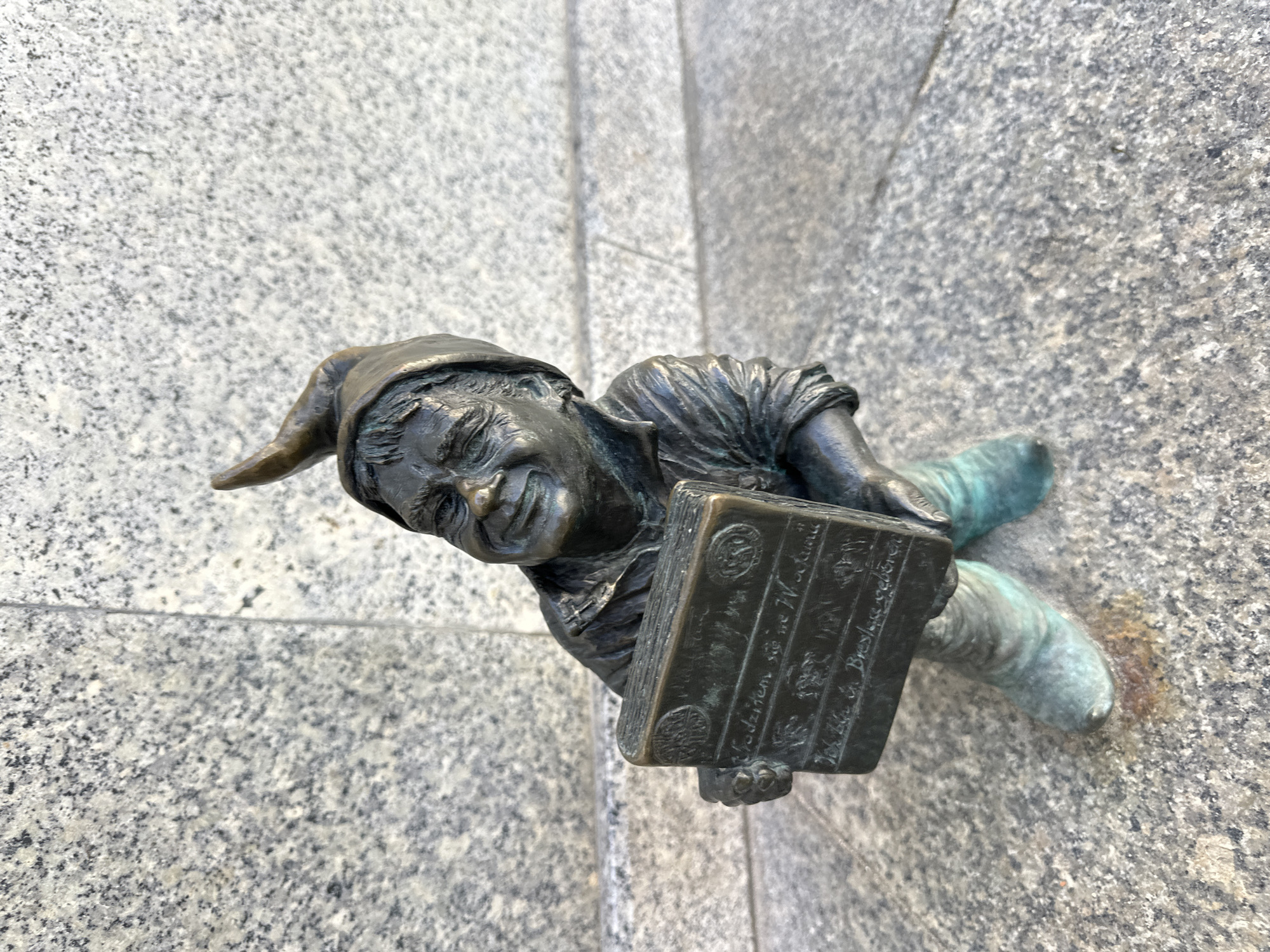 A close-up shot depicts a bronze dwarf statue affixed to a granite wall. The dwarf, wearing a pointed cap, holds a small, rectangular object resembling a book or tablet with illegible inscriptions. The statue exhibits a greenish-blue patina on the lower body, contrasting with the darker bronze of the upper body. The granite wall has a mottled gray and white pattern and a vertical seam is visible behind the statue. The statue's location is Wrocław, Poland, where such dwarf statues are a common urban feature.