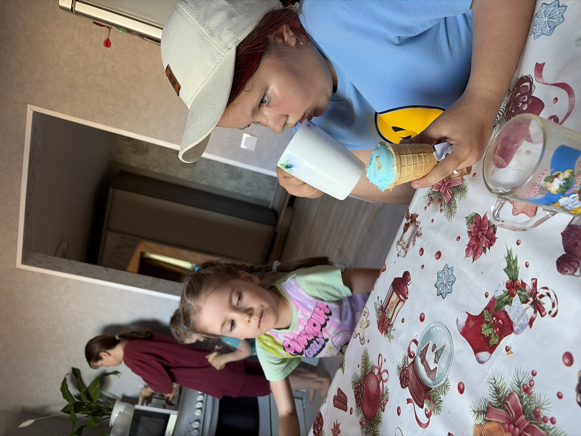 Two children are at a table covered with a Christmas-themed tablecloth. One child wears a white baseball cap and a blue shirt with a yellow smiley face, holding a cup in one hand and an ice cream cone in the other. The second child is wearing a green shirt with cartoon characters on it. In the background, an adult in a burgundy shirt stands near a stove with a potted plant next to it. The location is in a kitchen, possibly in Чумна, Russia.
