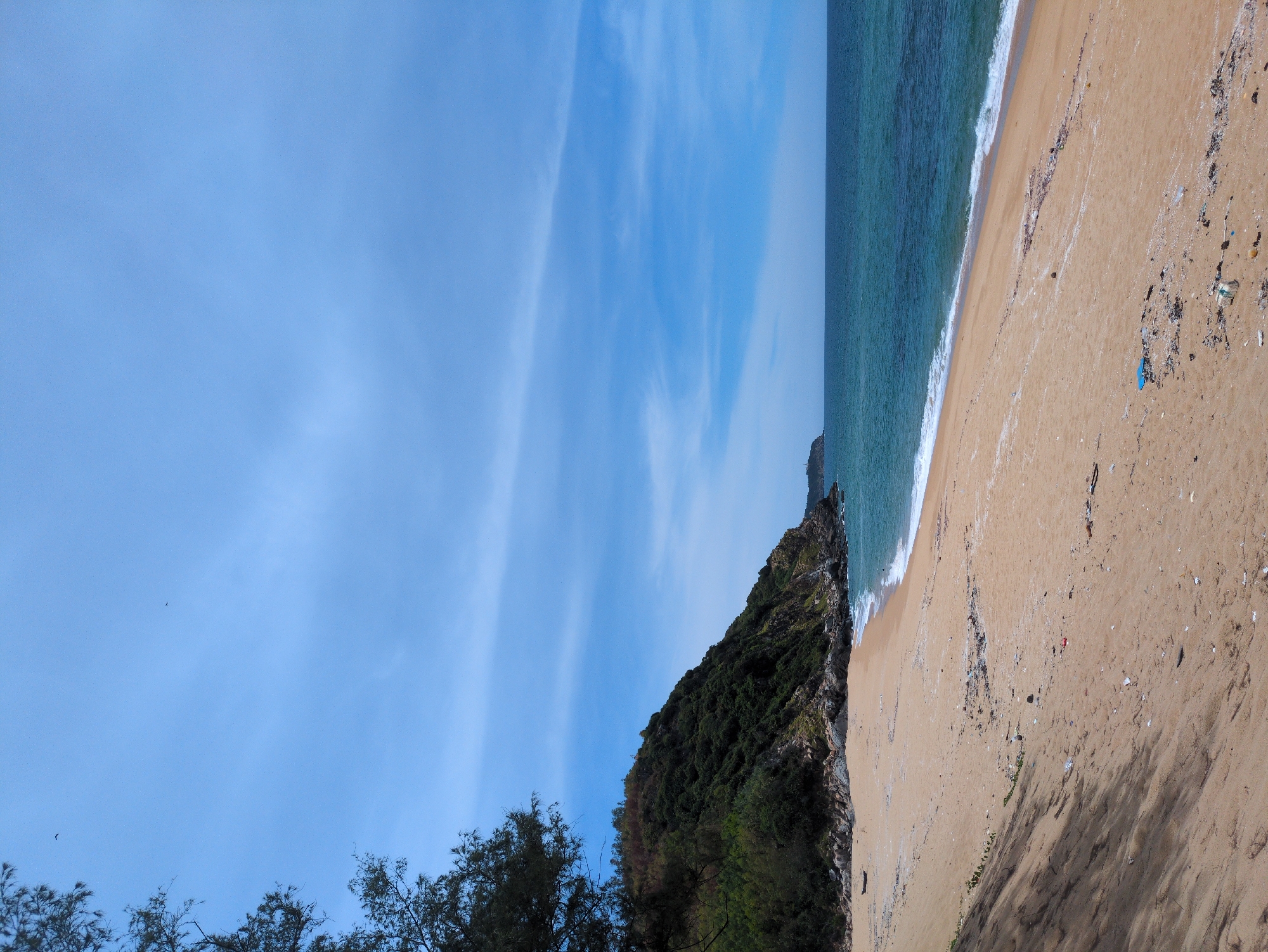 The image depicts a scenic view of a sandy beach in Sa Huynh Ward, Vietnam, stretching towards a rugged, tree-covered hillside on the left. The ocean, with its shades of blue and green, meets the shore with gentle waves. The sky is a clear blue, interrupted by thin, wispy clouds. The beach is mostly deserted, with some scattered debris, including plastic waste, visible on the sand. A lone, small boat is resting on the sand near the water's edge. The overall atmosphere is tranquil and natural, with a hint of human impact due to the presence of litter.