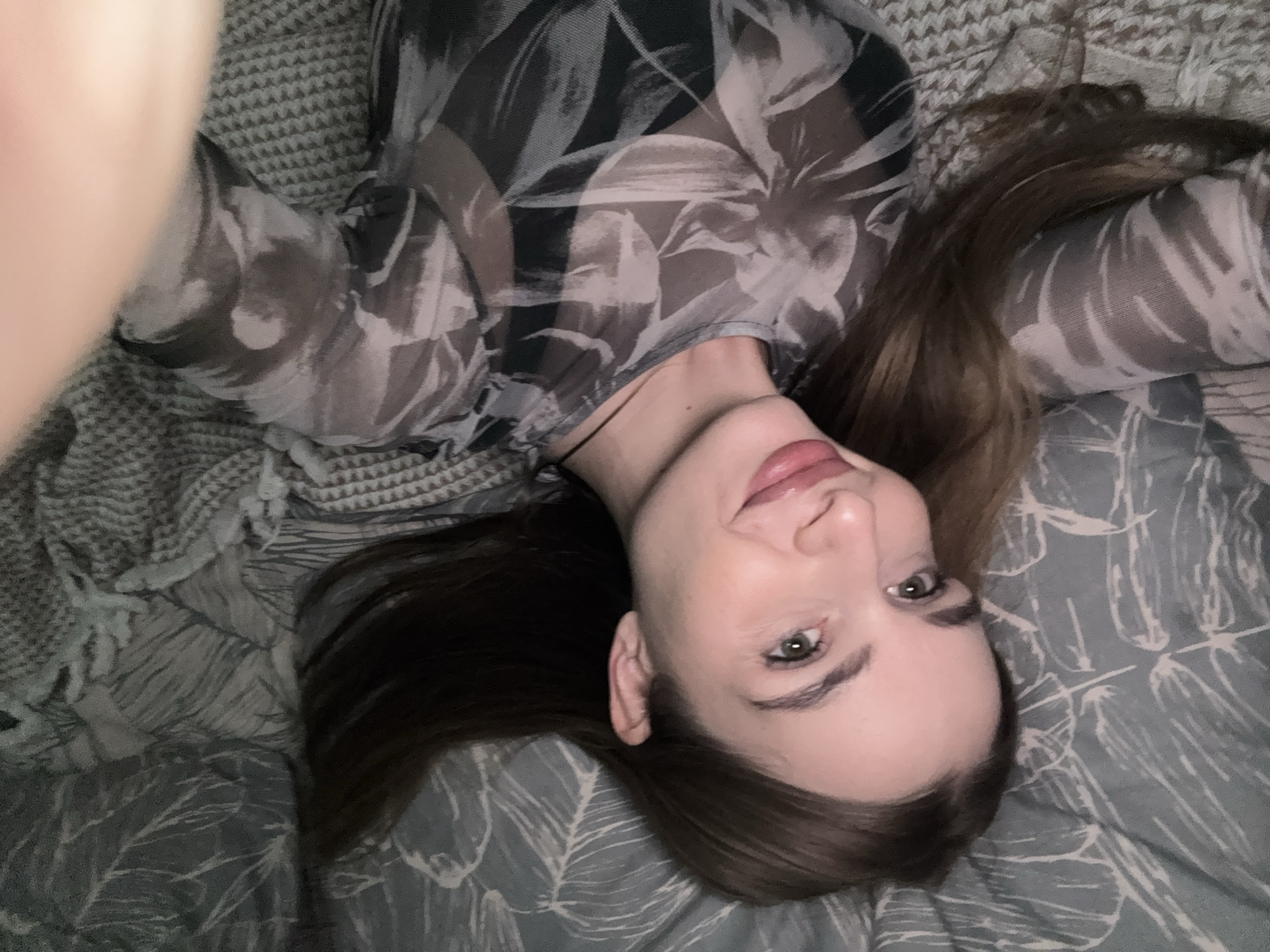 A woman with long brown hair is lying on a bed with patterned grey and white bedding. She is wearing a sheer top with a floral pattern. Her head is tilted towards the camera, and she is smiling slightly. The location is not discernible from the image.