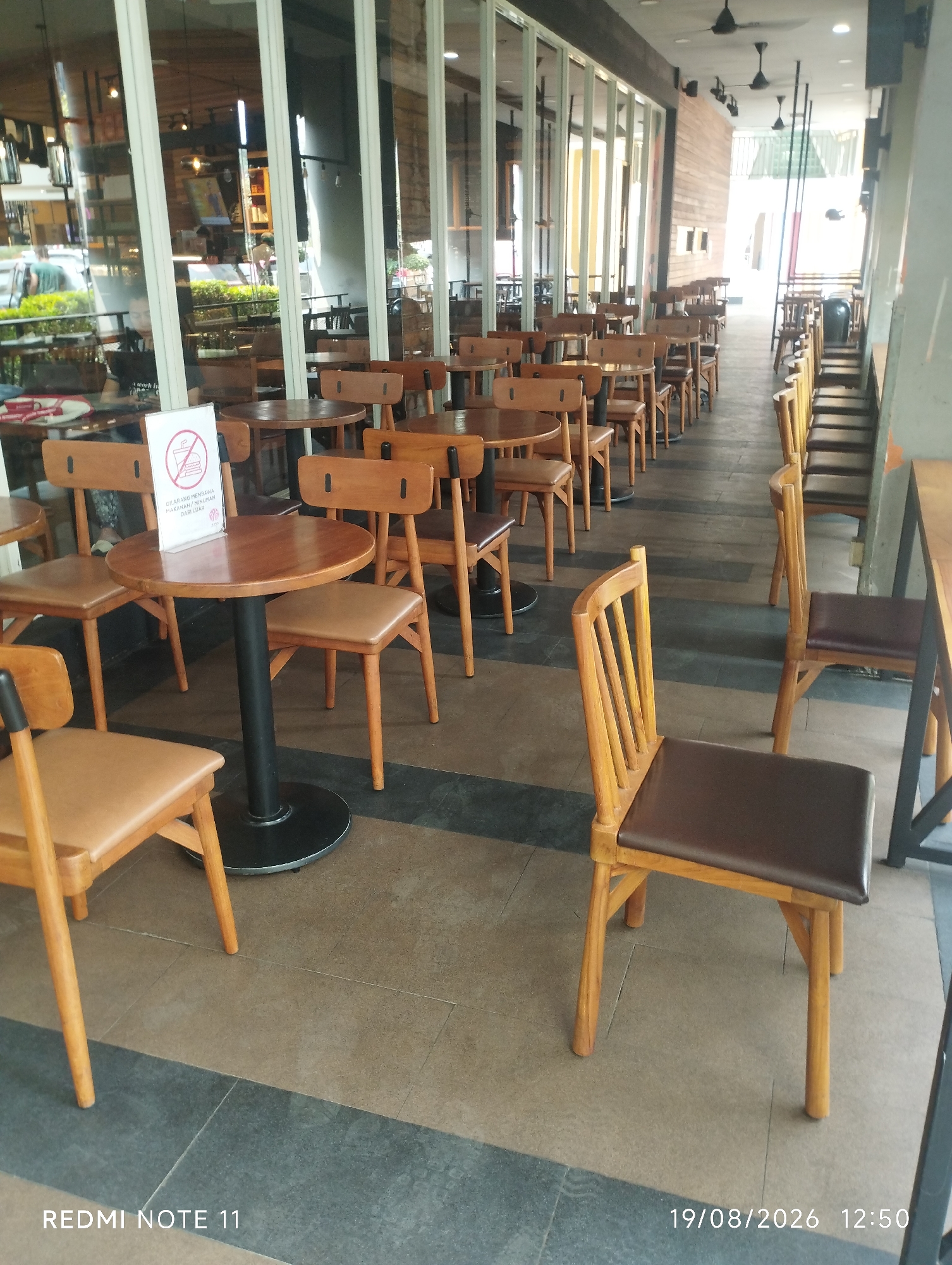 The image captures an outdoor seating area of a cafe or restaurant in Karawang, Indonesia, on August 19, 2026, at 12:50 PM. The scene is characterized by numerous wooden tables and chairs arranged on a tiled floor. The seating area is partially covered by a modern building with large glass windows and a wooden-paneled wall. In the foreground, a circular wooden table with a black metal base is surrounded by wooden chairs with brown leather seats. A white sign with a red circle and a line through a hamburger icon, indicating no food, is placed on the table. Further back, more tables and chairs are visible, some occupied by people, suggesting a dining or social setting. The environment appears to be clean and well-maintained, with natural light illuminating the area.