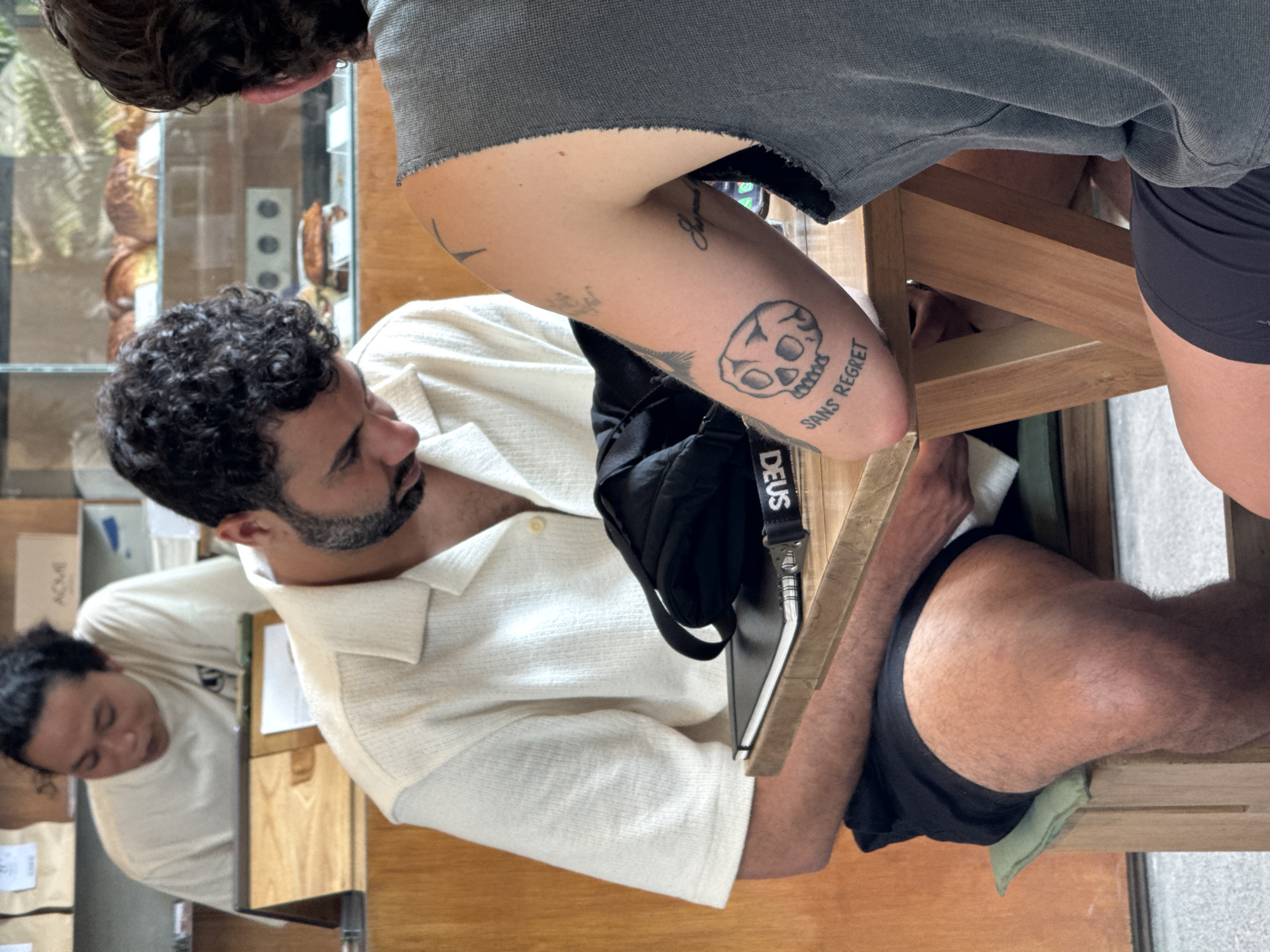 The image shows an indoor scene in what appears to be a cafe in North Kuta, Indonesia. Three people are present. The main subject is a man with dark curly hair and a beard wearing a white button-down shirt. He is seated at a wooden table, and a black bag with the word "DEUS" on the strap is placed near him. The top half of another person wearing a grey t-shirt and dark shorts stands near the main subject. This person has tattoos on their right arm including a skull and the words "Sans Regret." Partially visible in the foreground is a third person, likely a barista, with dark hair wearing a white t-shirt. They appear to be behind a counter. A glass display case with pastries and other baked goods is visible behind the main subject. The overall environment suggests a casual dining or cafe setting.