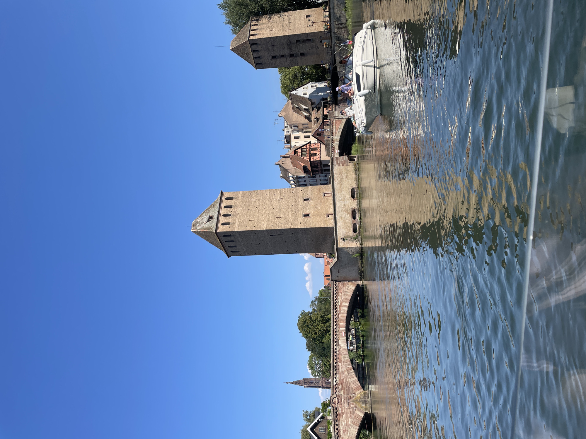 The image captures a view of the historic Ponts Couverts and the Vauban Dam in Strasbourg, France, on a clear, sunny day. The Ponts Couverts are a complex of three stone bridges, flanked by four tall defensive towers. These structures are made of reddish-brown stone, with arched openings.
In the foreground, a body of water, likely the River Ill, reflects the sky and the surrounding architecture. A white motorboat is visible in the middle right of the image, carrying at least three people who appear to be enjoying a leisurely ride. The boat has a dark canopy and is moving gently on the water.
In the background, beyond the Ponts Couverts, several buildings with characteristic Alsatian architecture can be seen. These buildings feature steeply pitched roofs and a mix of stone and timber-framed facades. A tall, slender spire of a church, likely part of the Strasbourg Cathedral complex, rises above the treeline on the left side of the image. Lush green trees are interspersed among the buildings, adding to the picturesque scenery. The sky is a vibrant, cloudless blue, indicating pleasant weather conditions. The overall scene depicts a scenic and tranquil urban landscape, characteristic of Strasbourg's historic quarter.
