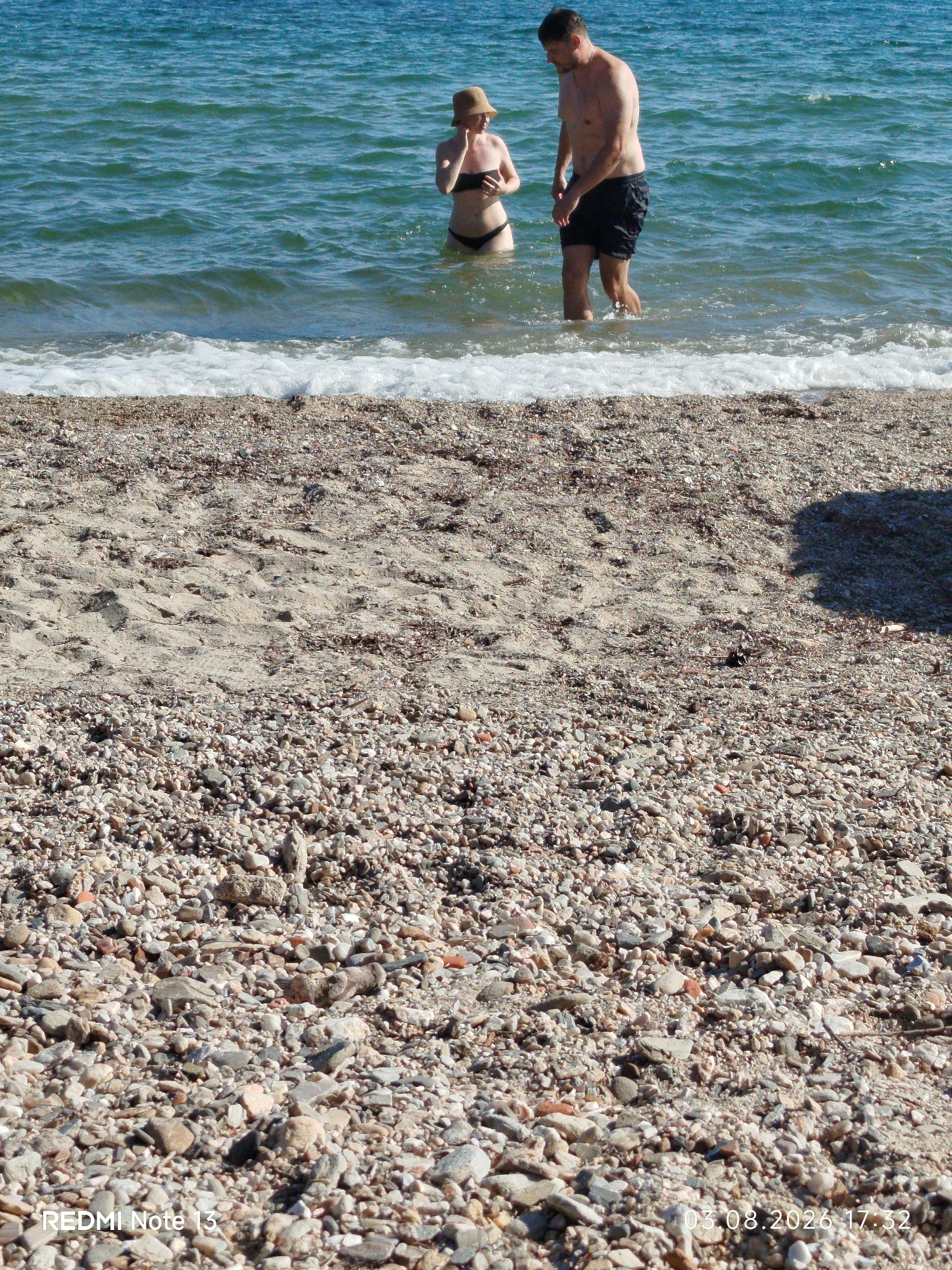 The image shows two people standing in the shallow water of a beach in Sithonia Municipal Unit, Greece. The person on the left is a female wearing a bikini and a hat. The person on the right is a male wearing black swim trunks. The beach is covered in pebbles and sand, and the sea is blue and calm. The date stamp indicates August 3, 2026, at 17:32.