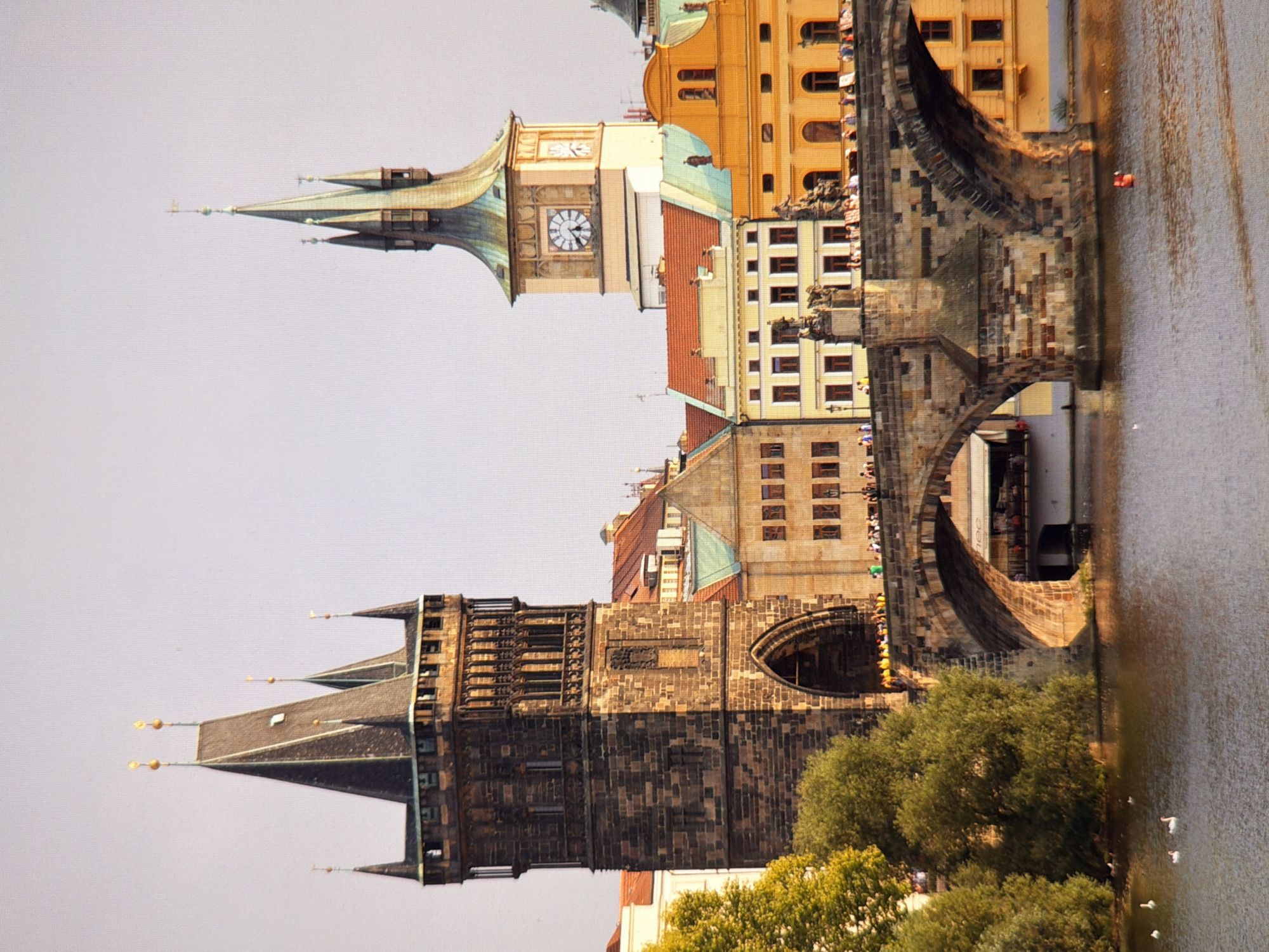 The image depicts a view of the Charles Bridge in Prague, Czech Republic. In the foreground, a body of water, likely the Vltava River, reflects the warm tones of the sunset. Several white waterfowl are visible on the water. The iconic Charles Bridge, a stone bridge adorned with statues, spans the river, and a procession of people are walking across it. The background features several historic buildings with red-tiled roofs, including the Old Town Bridge Tower on the left and a tower with a clock face on the right. Lush green trees, likely on the riverbank, frame the left side of the scene. The sky is clear and a pale, neutral color. There is no indication that this scene is located in Kyiv, Ukraine.