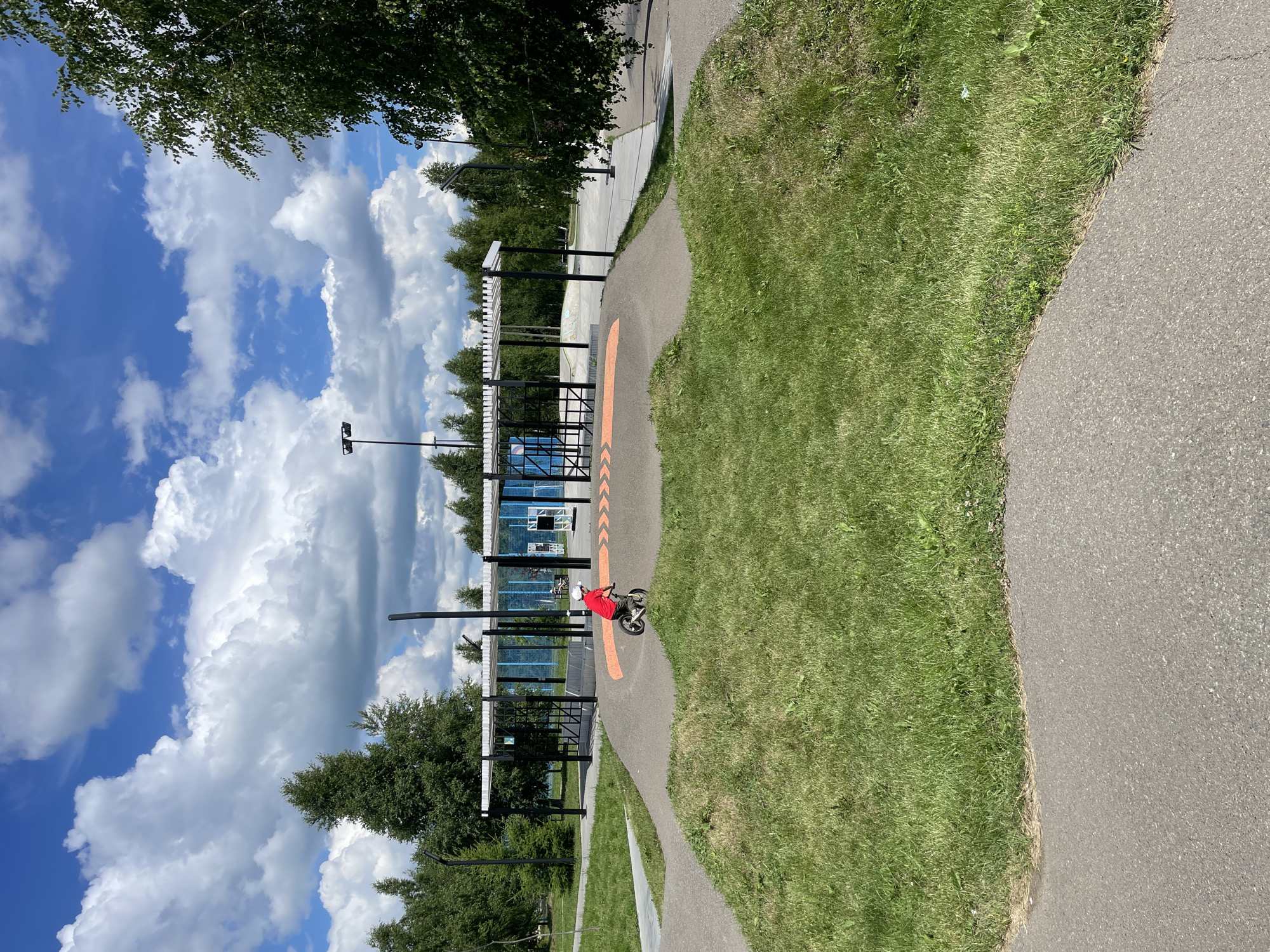 A boy in a red shirt rides a bicycle on a dirt track in a park. The track has orange arrows painted on it, indicating the direction of travel. In the background, there is a covered area with metal poles and a white roof, possibly a pavilion or shelter. Behind that, there are trees and a blue sky with white clouds. The scene appears to be a recreational area designed for cycling or other outdoor activities.