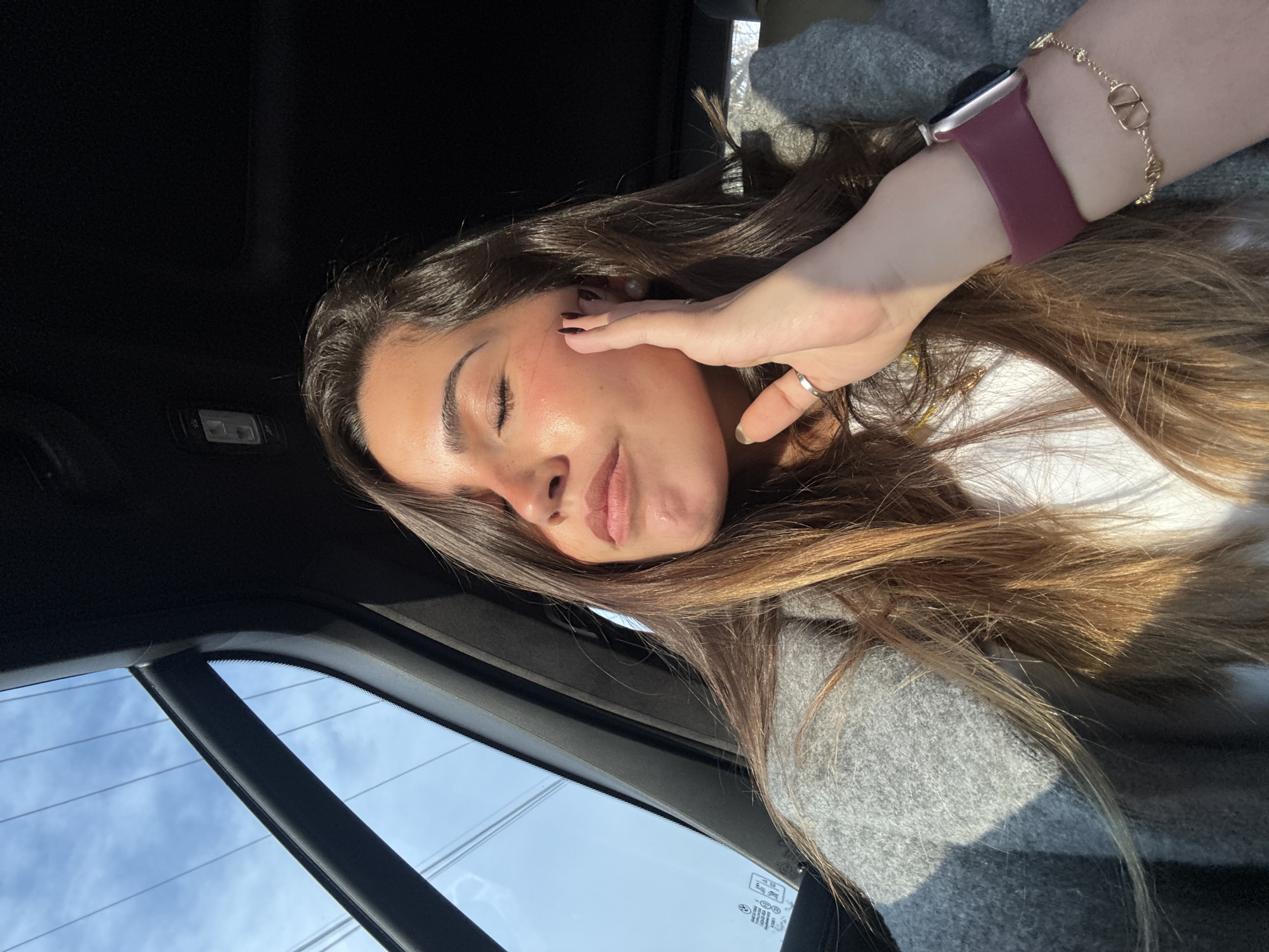 A young woman is reclining in a car. She has long brown hair and is wearing a light gray sweater over a white shirt. She is wearing a watch with a dark band and a gold bracelet on her wrist. Her eyes are closed, and her left hand is touching her face. The car window is visible in the lower left of the frame, showing a blue sky with utility wires. The interior of the car is black. There is no indication of the city of San Bernardino Tlaxcalancingo, Mexico.