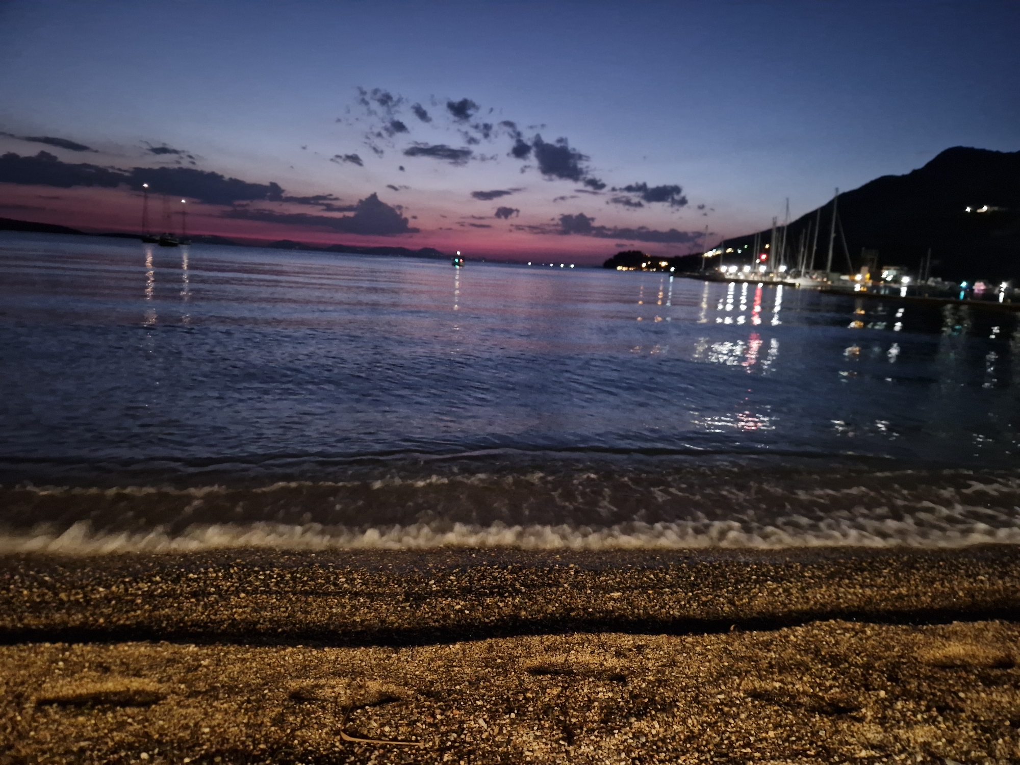 The image depicts a twilight scene at a beach, likely in Platariá, Greece. In the foreground, a pebbly beach meets the gentle waves of the sea. The water is dark and reflects the lights of a nearby town or harbor, which is visible in the middle ground. These lights shimmer on the water's surface. Further in the distance, silhouetted hills or mountains form the backdrop against a sky that transitions from deep blue to hues of pink and purple, indicating the last moments of sunset. Several sailboats are anchored in the bay, their masts visible against the fading light. The overall atmosphere is serene and peaceful.