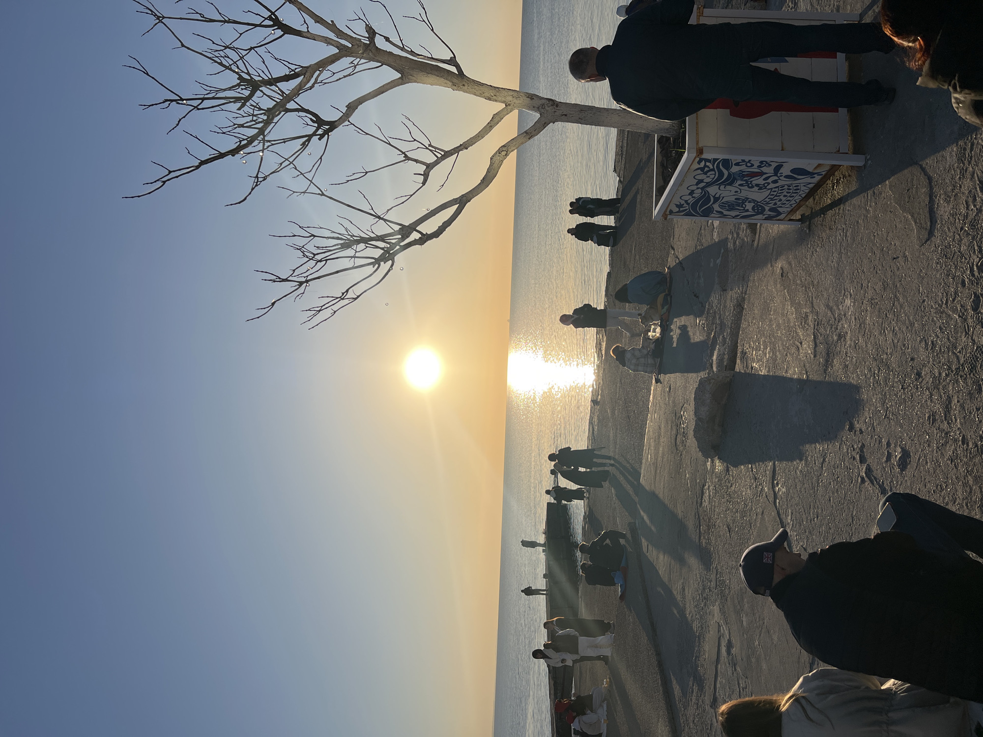 The image captures a waterfront scene during sunset in Unknown, Unknown. The sun's bright glare reflects off the water. A leafless tree is positioned on the left side of the frame. There are people gathered on a concrete pier or walkway. Some are standing near the edge of the water, while others are seated on the ground or on benches. Shadows cast by the people and the setting sun stretch across the concrete. One person appears to be standing on a decorated structure of some kind.