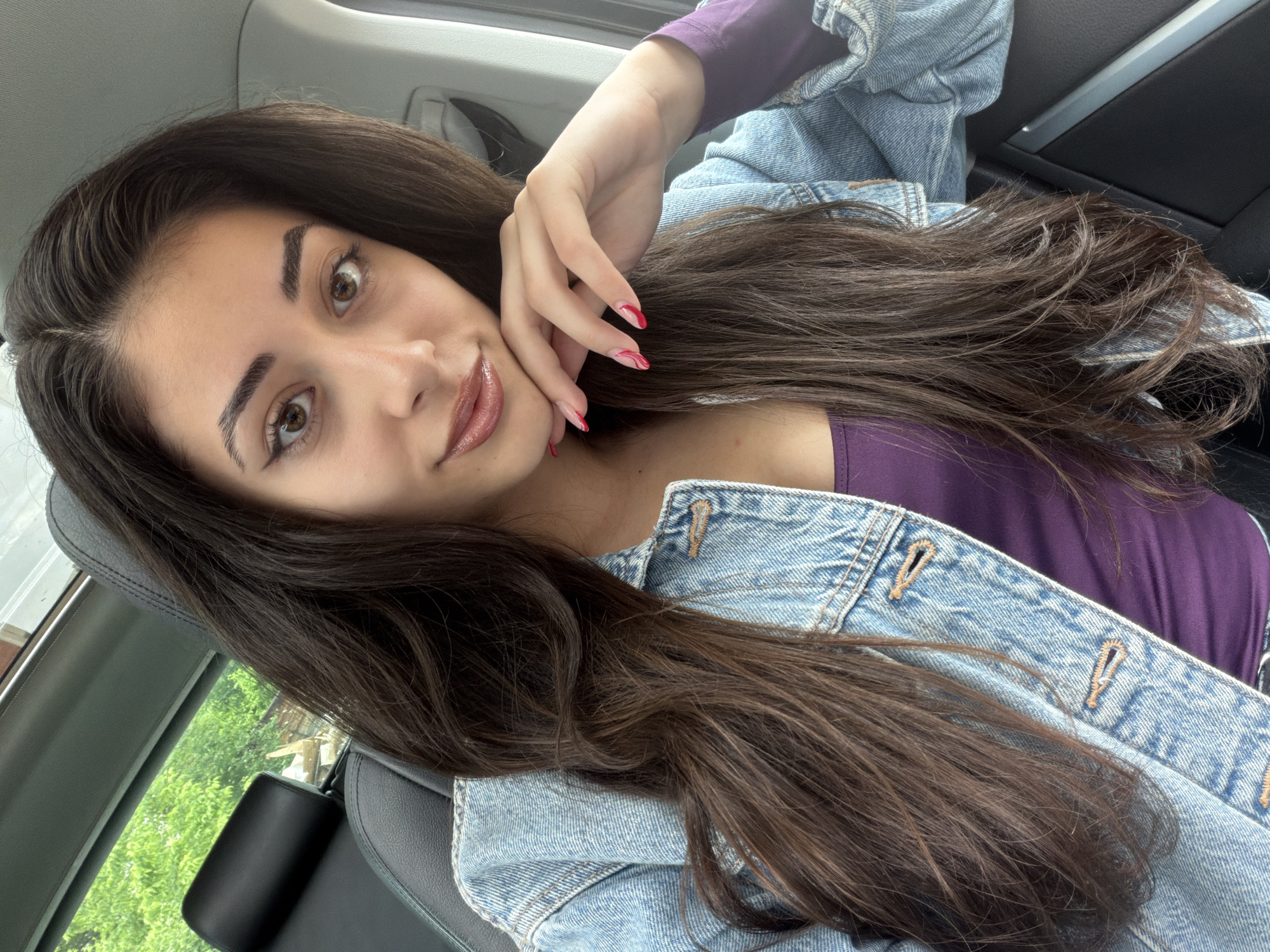 A young woman with long, dark brown hair is posing for a selfie inside a vehicle. She has light skin, brown eyes, and is wearing makeup that includes lip gloss. She is dressed in a purple top and a light blue denim jacket. Her left hand is raised near her face, with fingernails painted in red. The interior of the car is visible, including the black leather seat and parts of the car's interior trim. The lighting suggests it is daytime. The location in Sofia, Bulgaria cannot be confirmed from the image.