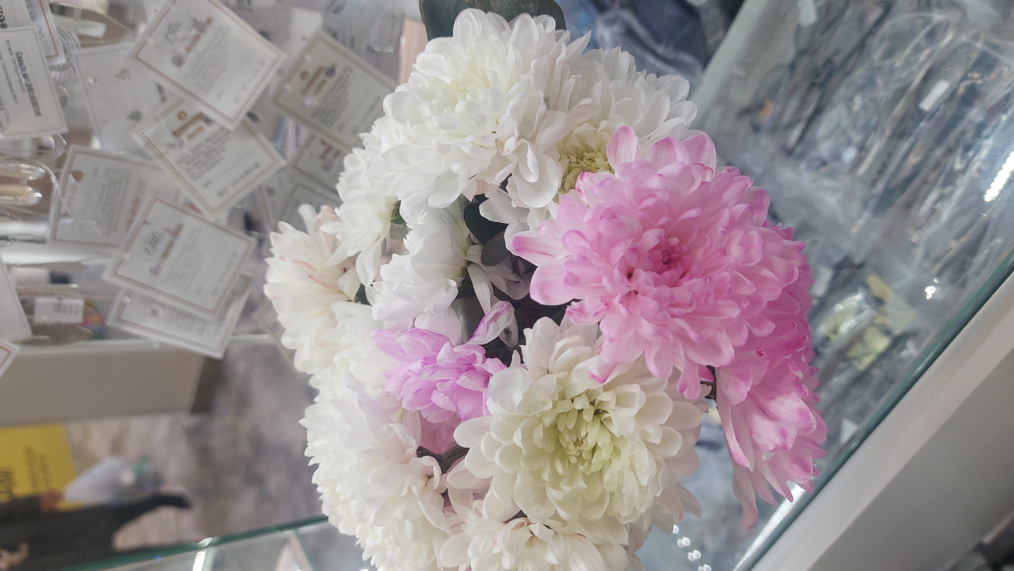 A still, close-up shot shows a bouquet of chrysanthemums, primarily white with some light pink, arranged in a glass display case. The bouquet is centered and slightly angled to the right. The background features a collection of small, rectangular tags or cards with text, possibly price tags or labels, scattered behind the bouquet. Some optical frames can be seen as well. The location appears to be a retail or commercial setting, potentially a shop in Novosibirsk, Russia. The lighting is somewhat diffused, creating soft shadows. The composition emphasizes the flowers and their textures, while the background provides context to the environment.