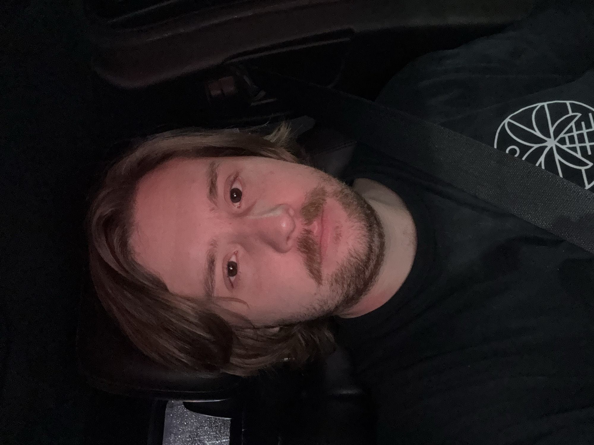 A medium-lit, low-angle close-up of a light-skinned male inside a vehicle. The man has shoulder-length light brown hair, light facial hair, and brown eyes. He is wearing a black t-shirt with a geometric design in white. A black seatbelt crosses over his torso. The setting is dark, suggesting nighttime. No indication of Mexico City.