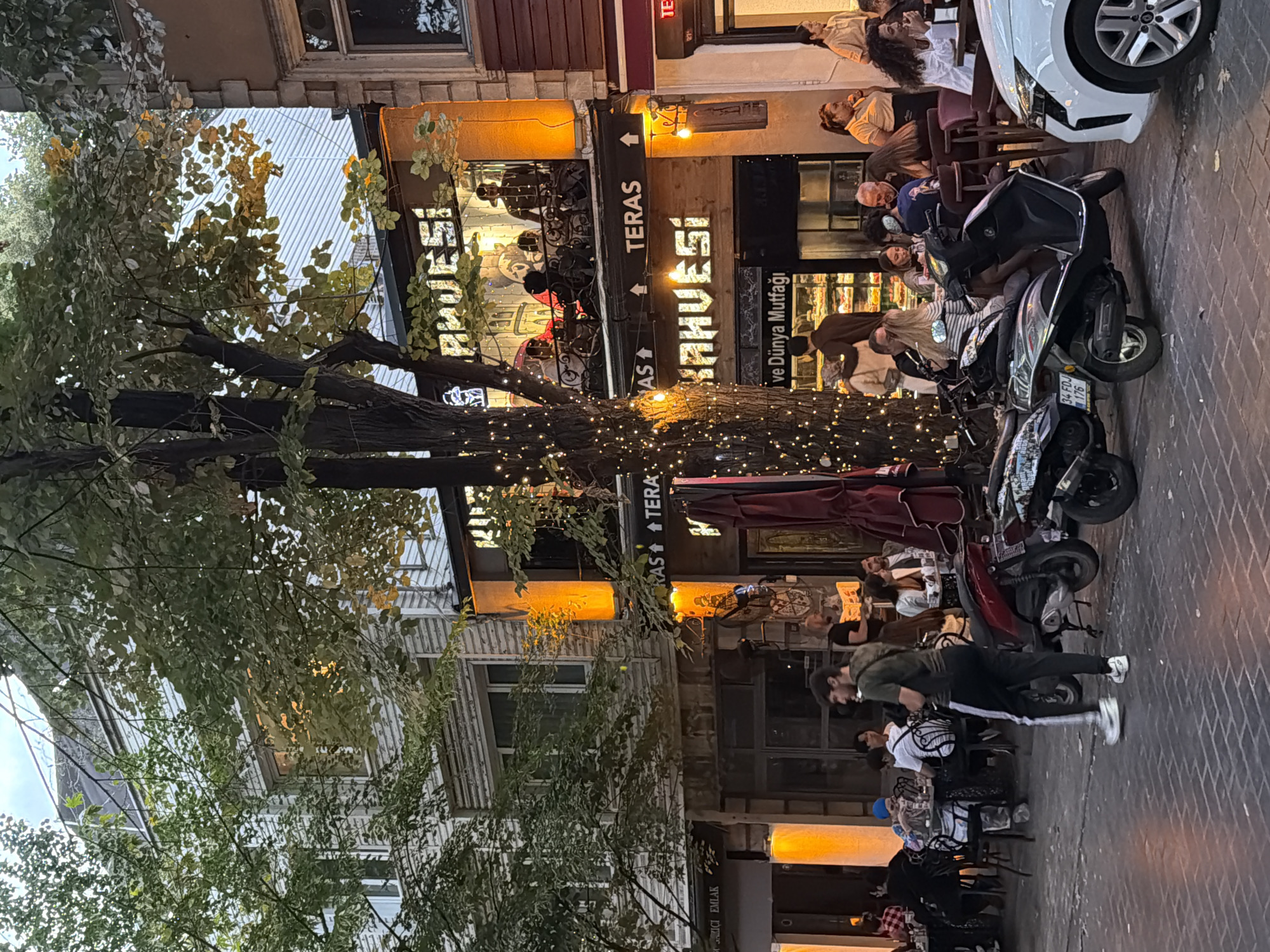 The image depicts a street scene in Üsküdar, Turkey, likely during the evening, as indicated by the artificial lighting. On the right side of the frame, a white car is partially visible. In front of a building with several floors, a ground-level establishment with tables and chairs is set up outdoors. A significant portion of the building facade displays signage with Turkish text, including "TERAS KAHVESİ". The second floor of the building features a balcony area where patrons are seated. Another establishment below the balcony has a display window showcasing food items.
In the foreground and midground, several people are present. On the left, a young man in athletic wear walks across the brick-paved street. Other individuals are seated at tables, some engaged in conversation and eating. Several scooters are parked along the sidewalk, one of which is prominently positioned in the center of the image. Lush green trees with lights strung around them partially obscure the view of the buildings. The overall atmosphere appears to be that of a lively urban dining and social area.