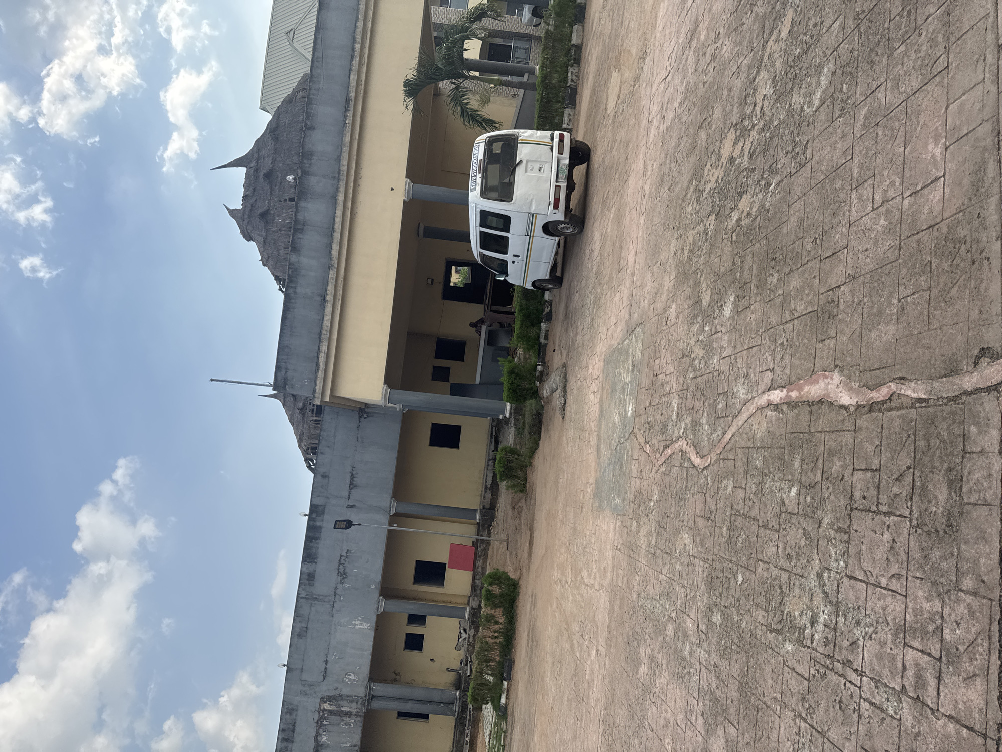 The image shows a partially dilapidated building in Ihiagwa, Nigeria. The building features a beige-colored main structure with gray concrete pillars and a traditional thatched roof element on top. A white van is parked in front of the building. The foreground is a concrete-paved area with visible cracks and wear. The sky is blue with scattered clouds. A palm tree is visible near the building.