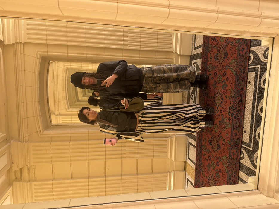 Two individuals are positioned in an ornate indoor hallway of a building in Paris, France, captured via a mirror reflection. The environment features light-colored, possibly cream, paneled walls adorned with classical pilasters and arched openings, creating an illusion of depth with receding reflections. The floor is composed of a dark and light geometric mosaic tile pattern, overlaid by a rectangular red oriental-style rug detailed with green, blue, and tan floral motifs.
The individual on the left, with dark, shoulder-length hair, wears a dark, possibly black, cropped jacket over a lighter top, a brown scarf, and wide-leg trousers with prominent black and white vertical stripes. She wears dark, platform-style footwear. She carries a large, light-colored tote bag with vertical stripes on her left shoulder and a smaller, olive-green hobo bag on her right. In her right hand, she holds a pink reusable cup with a dark logo, and in her left, a smartphone.
The individual on the right, featuring dark dreadlocks and a black beanie, is dressed in a dark, possibly black, padded jacket and baggy camouflage-patterned trousers. He wears dark, chunky boots. He holds a smartphone in his right hand, pointed towards the reflection, suggesting he is operating the camera. Both individuals stand side-by-side on the rug, directly facing their reflections.
