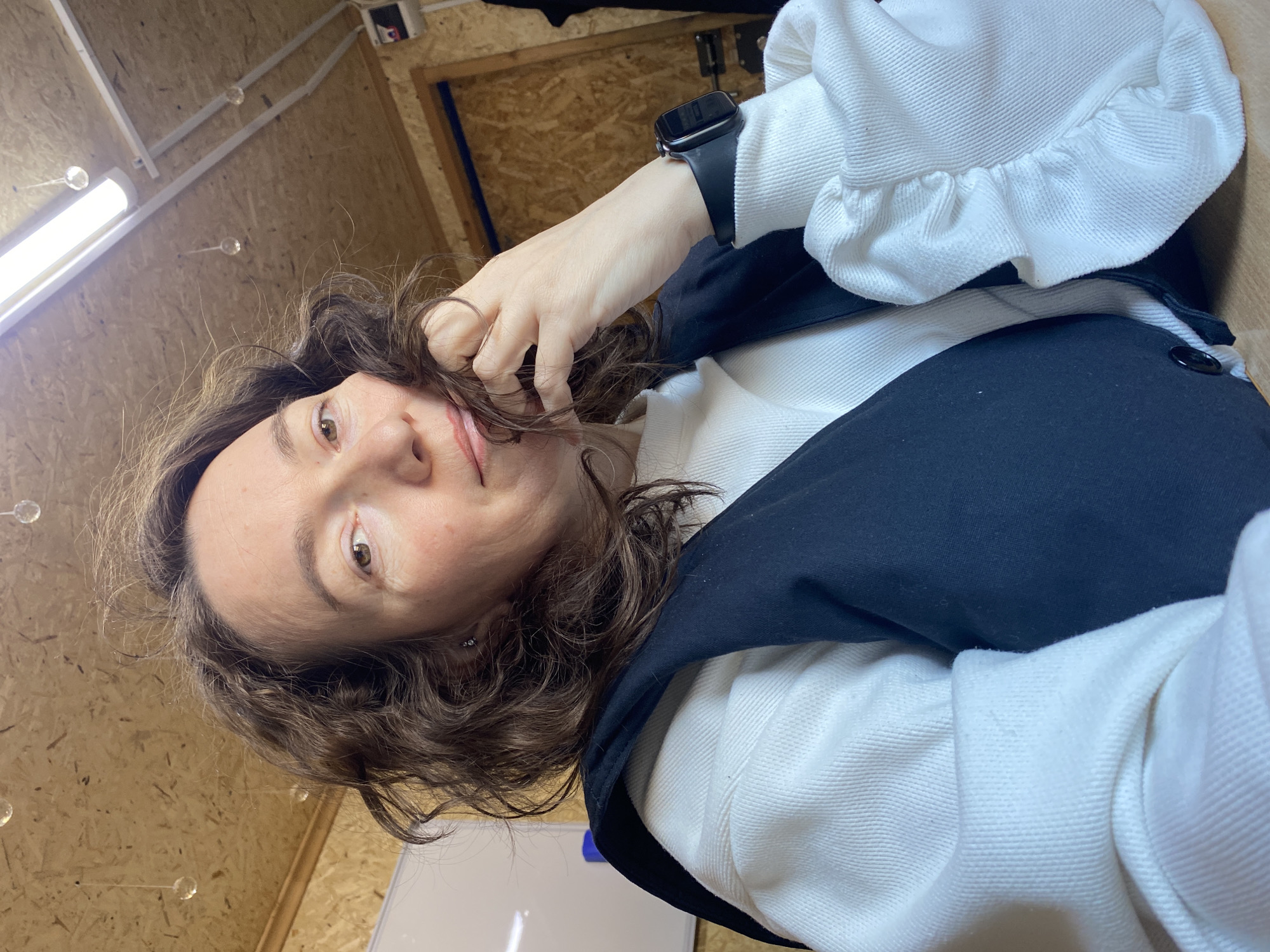 A selfie taken indoors. The subject is a woman with brown curly hair, wearing a white top with ruffled sleeves and a navy blue vest. She is looking directly at the camera, with one hand touching her hair. A smart watch is visible on her wrist. The background shows a wall constructed from light-colored wooden panels, and a fluorescent light fixture is partially visible in the upper left corner. A whiteboard is seen at the lower part of the image. The location is potentially a studio or workshop space, potentially Ol'khovskoye, Russia.