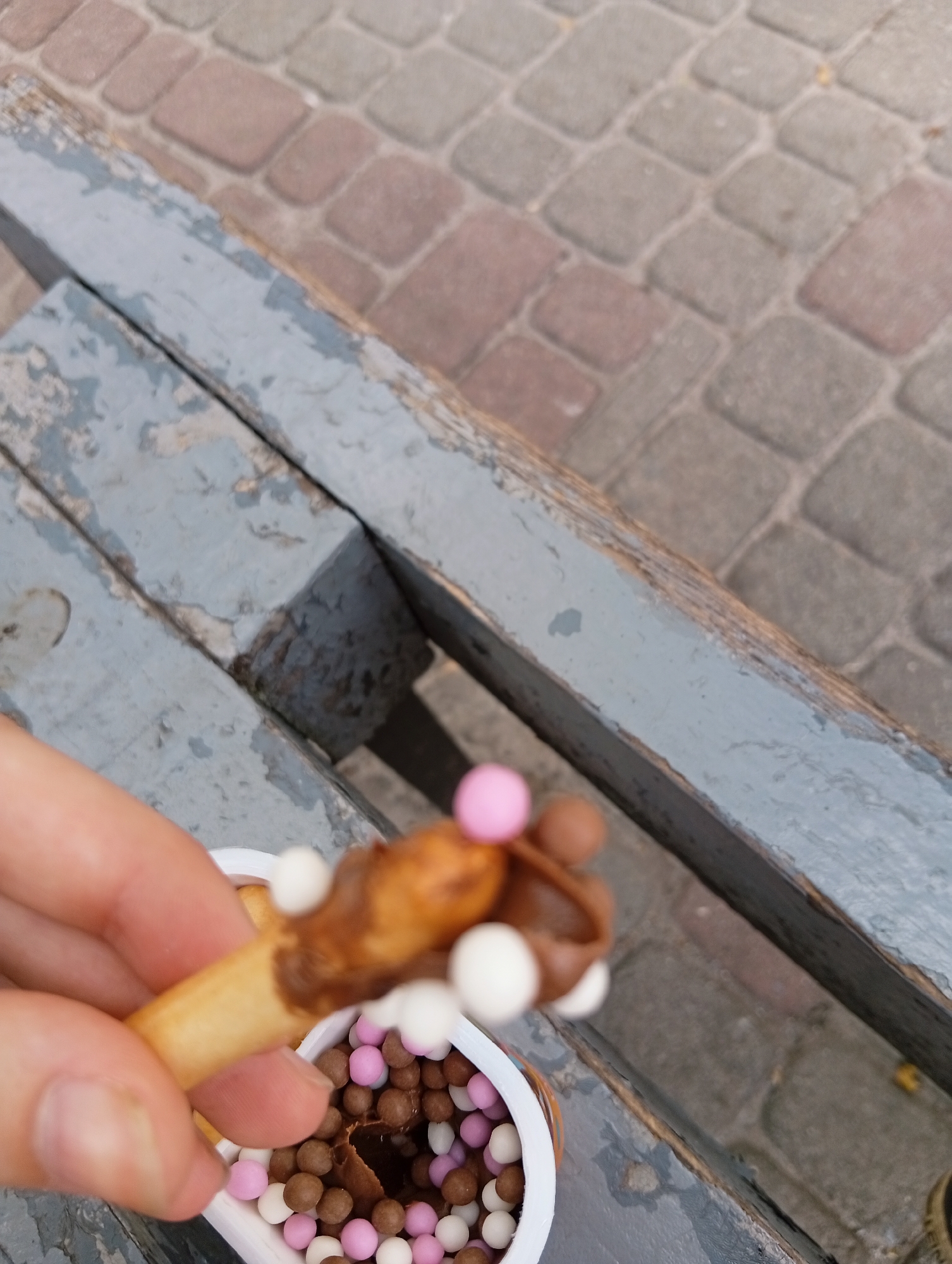 A light-skinned hand, likely an adult's, is holding a snack consisting of a light brown biscuit stick partially covered in dark brown creamy spread, adorned with small, spherical candies in white, light pink, and light brown. The snack is positioned above a white plastic container, which also holds the dark spread and an abundance of the same multi-colored candies, some of which are visible at the bottom of the container. A scoop mark is present in the spread within the container. The scene is set outdoors, with a weathered wooden bench in a faded, peeling blue-grey paint occupying the mid-ground. The planks of the bench are horizontal, showing significant wear and exposed wood. The background features paved ground composed of multi-toned grey and reddish-brown rectangular pavers arranged in a repeating pattern. The location is the city of Poltava, Ukraine.