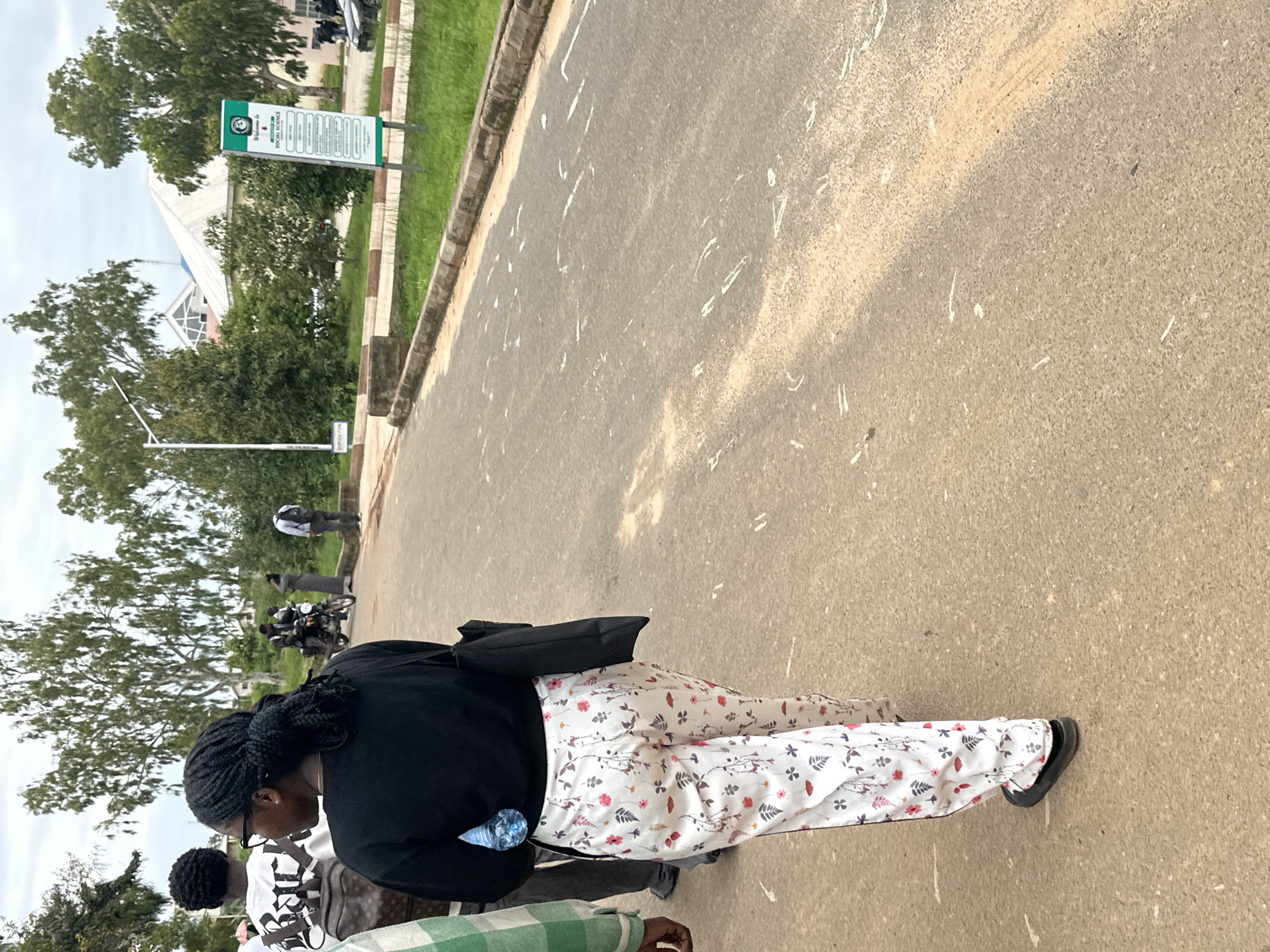 The image shows an outdoor scene on a paved road in Rafin Par, Nigeria. In the foreground, a young Black woman with braided hair and glasses is walking away from the camera. She is wearing a black long-sleeved top and white pants with a floral pattern. She is carrying a black tote bag over her right shoulder and a blue water bottle in her left hand. To her left, another person wearing a green and white checkered shirt and grey pants is partially visible.
Further down the road, several other individuals and objects are present. A person is riding a motorcycle, with another person standing behind them. Further in the background, there are buildings with white roofs surrounded by lush green trees. A green and white sign with text is visible on the right side of the road. The road itself is asphalt with some white markings and debris scattered on its surface. The sky is overcast.