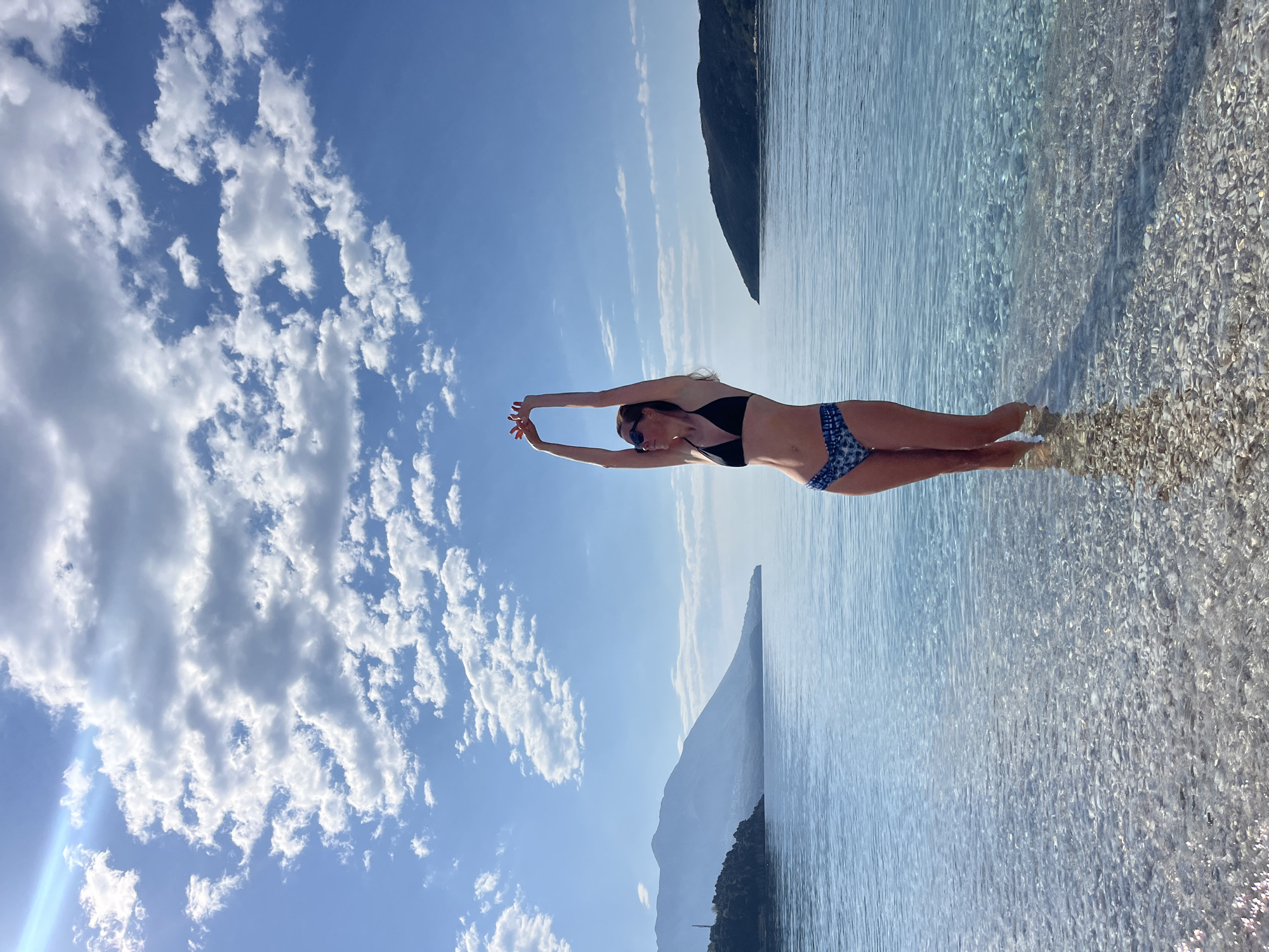 The photograph shows a woman standing in shallow, clear water on a pebble beach. She is wearing a black bikini top and blue patterned bikini bottoms, with her arms raised above her head. The sky is blue with scattered white clouds. In the background, there are landforms that appear to be mountains or hills rising from the sea. The location is Budva, Montenegro, based on the landscape and beach characteristics. The scene depicts a tranquil beach setting with a focus on the woman's pose.