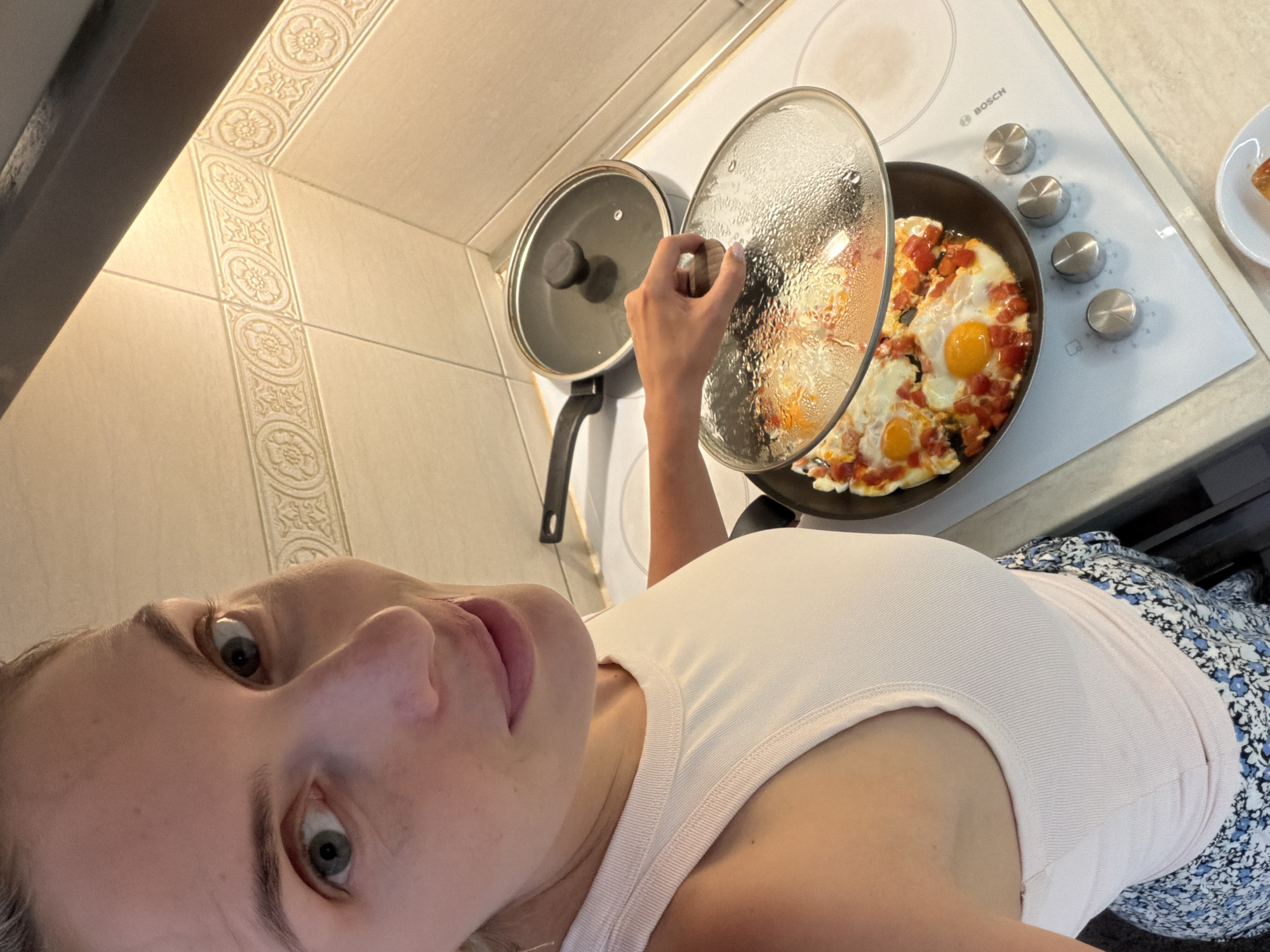 The image shows a woman in a kitchen, preparing a meal. She is wearing a white tank top and a floral skirt. In the foreground, the woman's face is partially visible, with her eyes wide open and a slight smile. She is looking towards the camera. In the background, a frying pan with eggs and chopped tomatoes is on a white Bosch electric stovetop. The woman's hand is lifting the glass lid of the pan, revealing the cooking food. The kitchen appears to have beige tiled walls with decorative borders. The overall lighting suggests it could be daytime. The woman's expression seems to indicate surprise or amusement. No visible text is present in the image, apart from the brand name "BOSCH" on the stovetop. The location context is Budva, Montenegro.