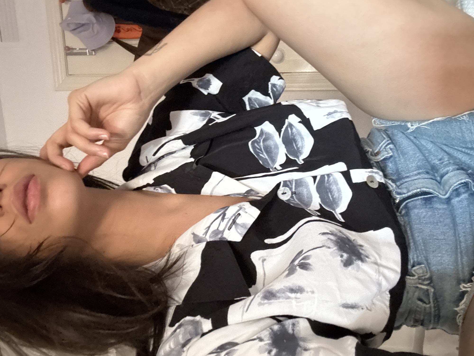 The media file is a close-up, low-angle, full-body shot of a woman lying down. She is wearing a black and white floral print button-up shirt and ripped denim shorts. Her legs are crossed, and her left hand is touching her face, near her mouth. The woman has dark hair, and appears to be resting or posing. The background is somewhat blurred, showing a wall, a mirror reflecting a hat, and some clothing items. There is no visible text in the image. The lighting suggests an indoor setting, possibly a bedroom, during the daytime. There are no clear indicators of the specific time of day or weather conditions. The overall mood is relaxed and casual.