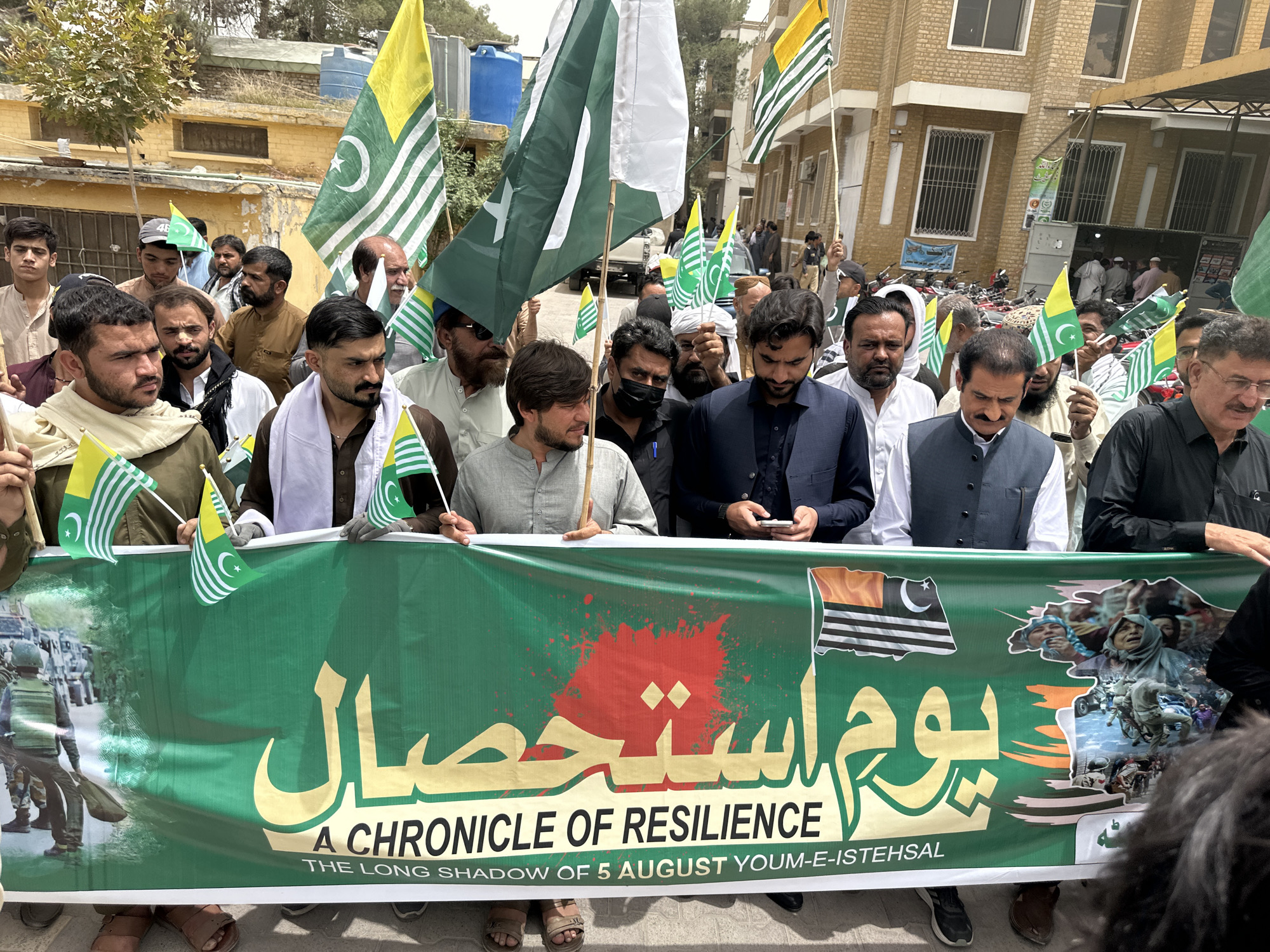 A group of men are holding flags and a banner in front of a building in Quetta City Tehsil, Pakistan. The banner is green with gold lettering and images of people and military vehicles. Several men are holding small flags that are green and white with a crescent moon and star. The building in the background is a light brown color with windows and a covered walkway. The men are dressed in a variety of clothing, including traditional Pakistani attire and more casual wear. The overall scene suggests a protest or demonstration.