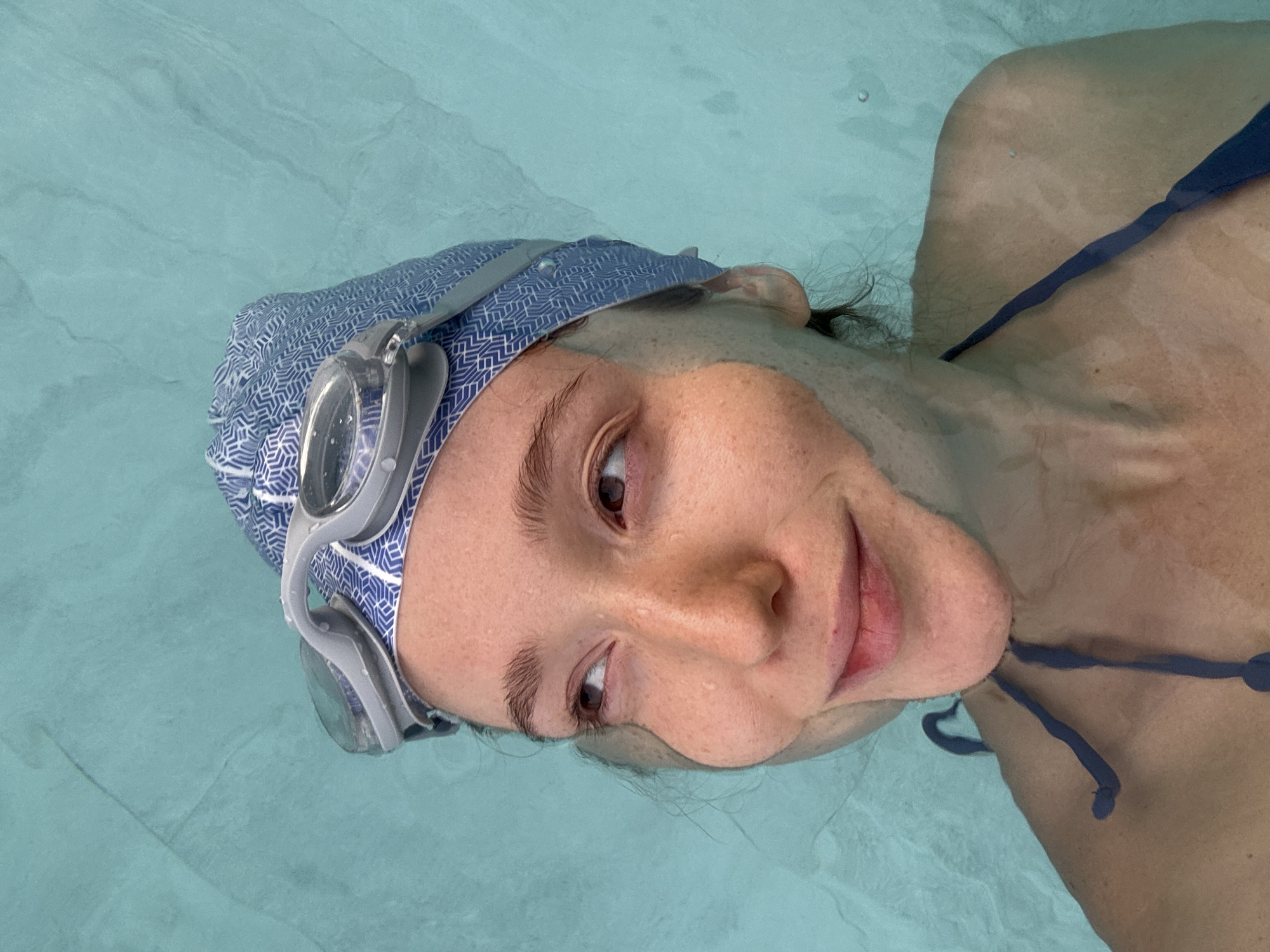 A person is captured in a close-up, slightly rotated view, partially submerged in clear, light blue water, likely a swimming pool in Choeng Thale, Thailand. The subject, appearing to be a woman, is wearing a blue and white patterned swim cap and clear swimming goggles. Her face, from her forehead to her chin, is visible above and through the water's surface, creating a distinctive distortion effect on her features below the waterline.
Her eyes are open and looking towards the upper right of the frame, with a subtle, relaxed smile gracing her lips, conveying a sense of calm enjoyment. Small water droplets are visible on her face. Dark hair strands are visible near her right ear, peeking from under the swim cap. A blue swimsuit strap is visible on her right shoulder and upper chest, submerged and appearing slightly blurred by the water. The water itself is serene, with gentle ripples and reflections on its surface. The overall impression is of a leisure activity, possibly swimming or floating, in a bright, tranquil setting. No text is visible in the image.