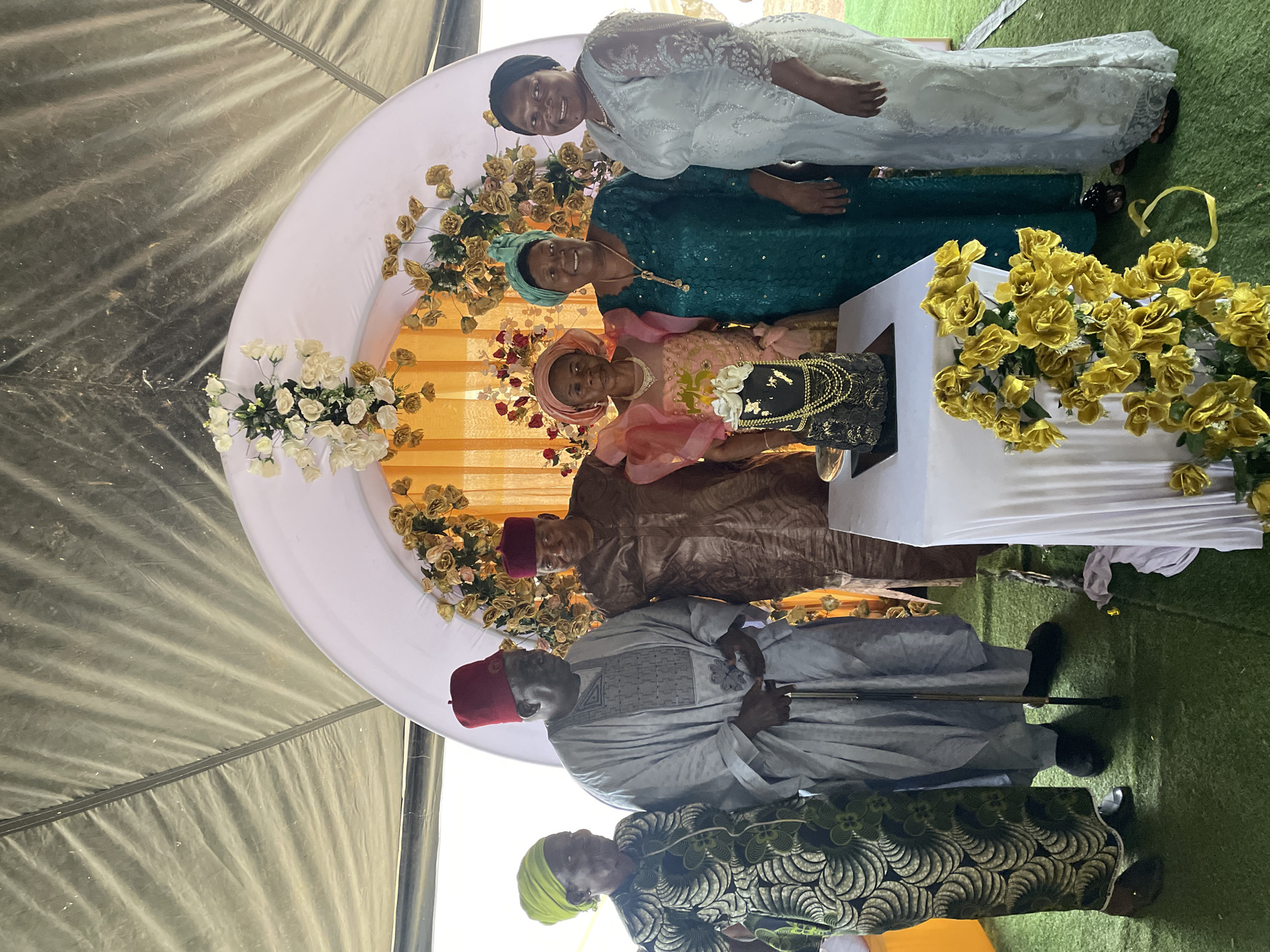 The photograph captures a group of seven people posing for a photo in what appears to be an event setting, possibly a wedding or a special celebration, within the city of Jos, Nigeria. The backdrop includes a semi-circular floral arrangement of white and yellow roses.
In the front row, a man with a red fez is holding a cane. Next to him is a woman wearing a green and black patterned dress.
The middle row includes a man in a brown robe with a red fez, and a woman in a pink outfit who is holding a black purse with a decorative gold handle.
The back row shows a woman wearing a green dress, and another woman in a white lace dress. They are all smiling, suggesting a joyous occasion. Yellow roses are clustered on the right. The scene takes place under the cover of a tent.