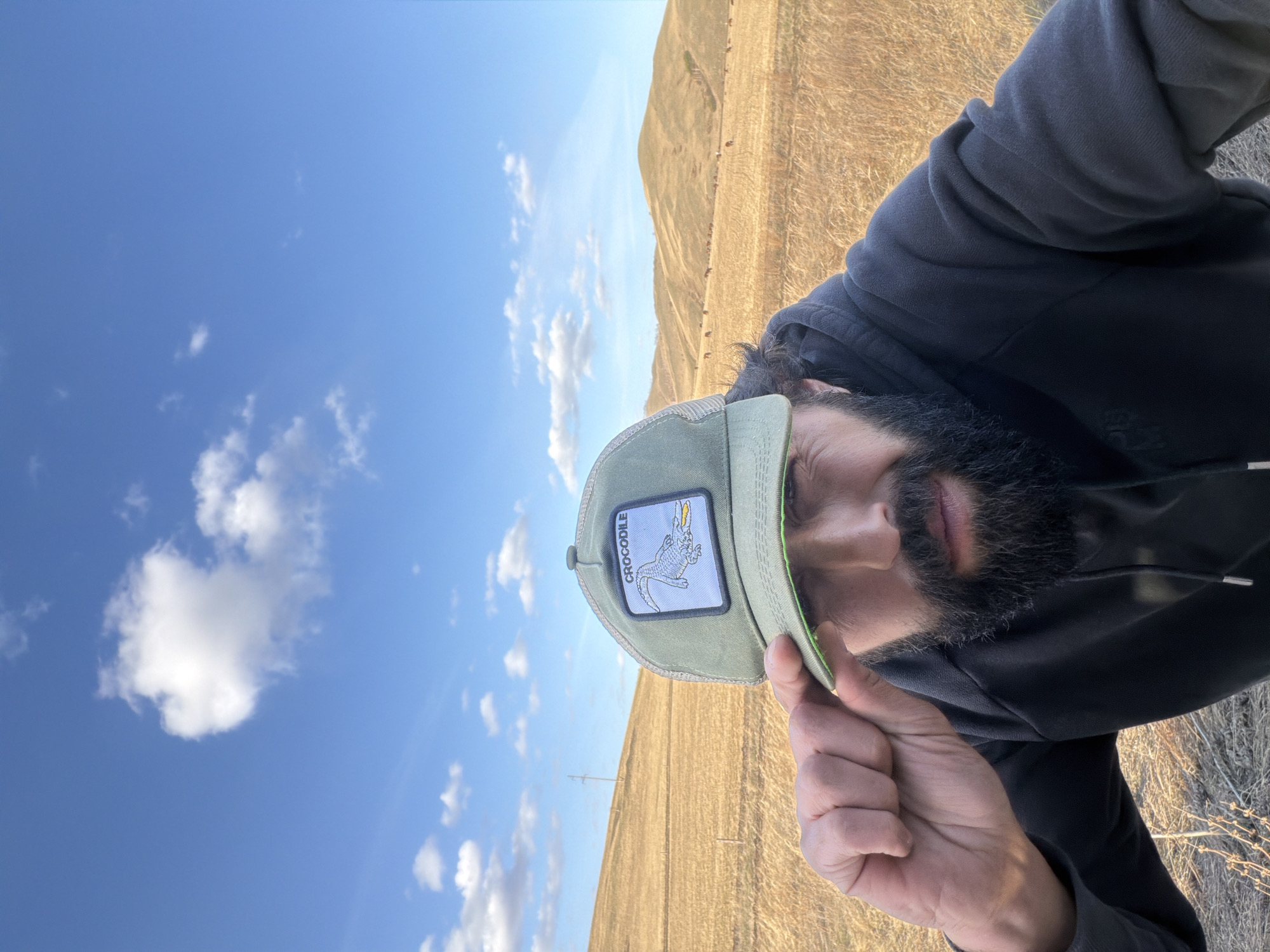 The image is a selfie of a person wearing a green cap with a crocodile emblem. The person is wearing a black hooded sweatshirt. The background consists of a blue sky with white clouds and a dry, grassy landscape. The image is taken outdoors. The location is the city of Первомайское, Russia.