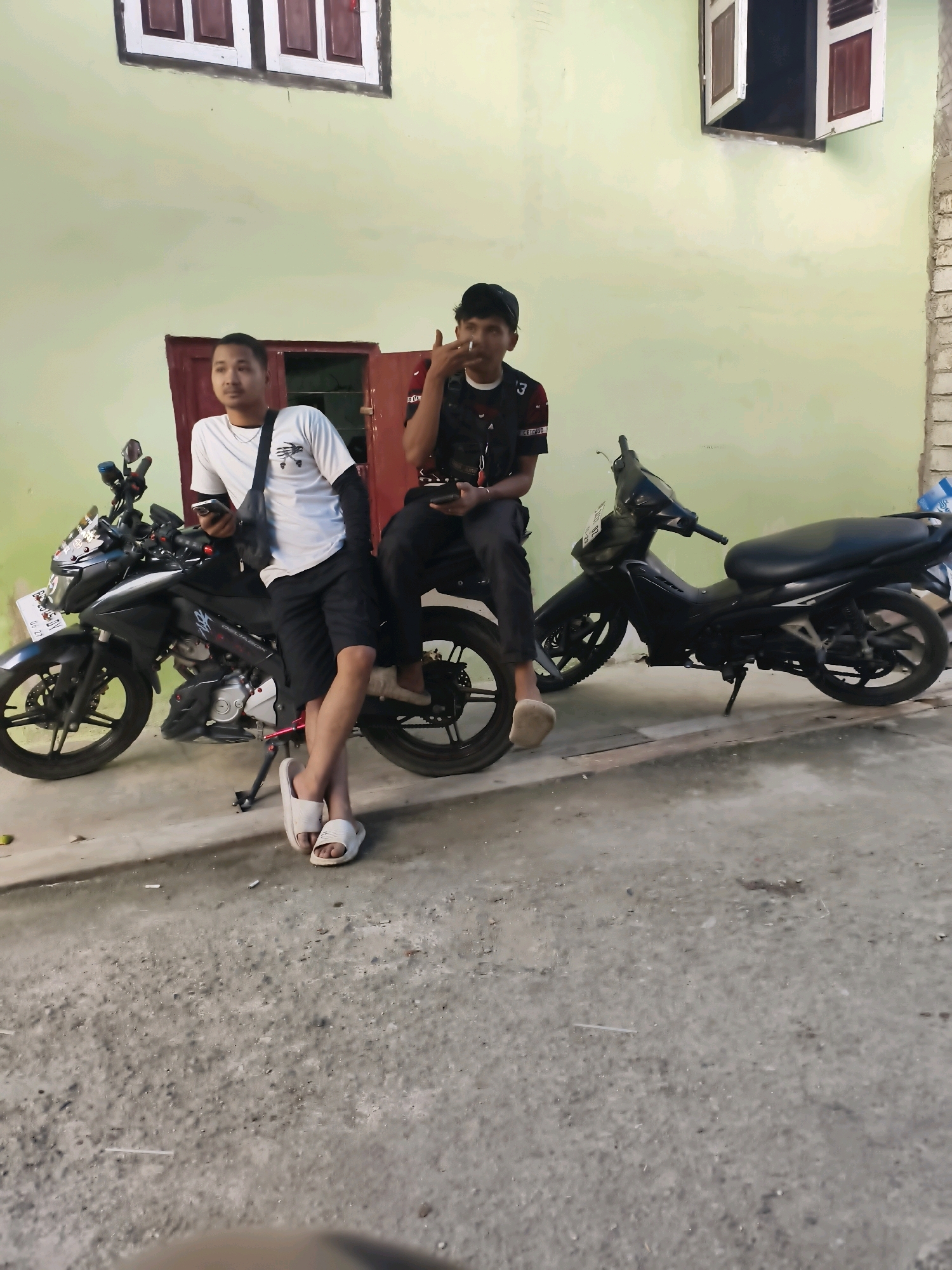 Two young men are standing or sitting near two motorcycles parked on a paved area in front of a light green building with red-trimmed windows. The man on the left, wearing a white t-shirt and black shorts, is leaning against a black motorcycle and holding a smartphone. He is wearing white sandals. The man on the right, wearing a dark t-shirt with a red stripe and a black baseball cap, is sitting on a motorcycle and smoking a cigarette. He is also holding a smartphone. Both motorcycles are facing to the left. The building appears to be a residential or commercial establishment. The scene is likely located in an urban or semi-urban area, possibly a street or alleyway in Sungai Penuh, Indonesia.