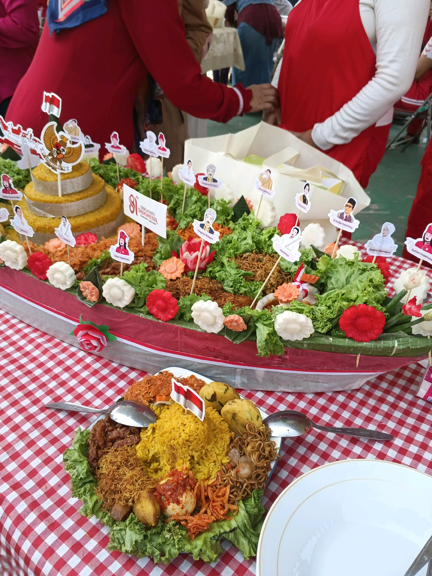 A large, boat-shaped dish filled with yellow rice, shredded chicken, noodles, hard-boiled eggs, and vegetables is displayed on a red and white checkered tablecloth. Several small Indonesian flags are stuck in the rice. In the background, there is a tiered, cake-like food structure decorated with small flags featuring images of people and an eagle. The entire scene appears to be set up for a celebration, possibly in an outdoor or semi-outdoor area, indicated by the presence of people in casual attire and a hint of greenery in the background. The food presentation suggests a cultural event.