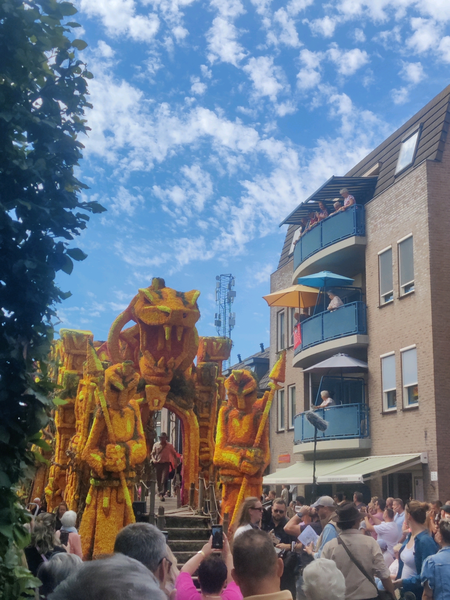 In Zundert, Netherlands, a vibrant street scene unfolds, featuring a parade float adorned with thousands of orange and yellow flowers. The float depicts a colossal, snarling creature resembling a tiger or dragon, with sharp teeth and menacing eyes, flanked by two warrior-like figures, also constructed from flowers. These figures stand guard, one holding a spear. The float is highly detailed and appears to be the central attraction of the event.
A crowd of spectators lines the street, observing the parade. Many are holding up their phones to capture photographs and videos of the spectacle. The people in the foreground are varied in age and attire, some wearing casual summer clothing. Behind the float and the crowd, a multi-story brick building stands. Several balconies on this building are occupied by people watching the parade from above. Umbrellas, both open and closed, are visible on the balconies, suggesting sunny weather. The sky above is a bright blue, dotted with fluffy white clouds. The overall atmosphere suggests a festive outdoor event, likely a flower parade given the context of Zundert, known for its annual flower parade.