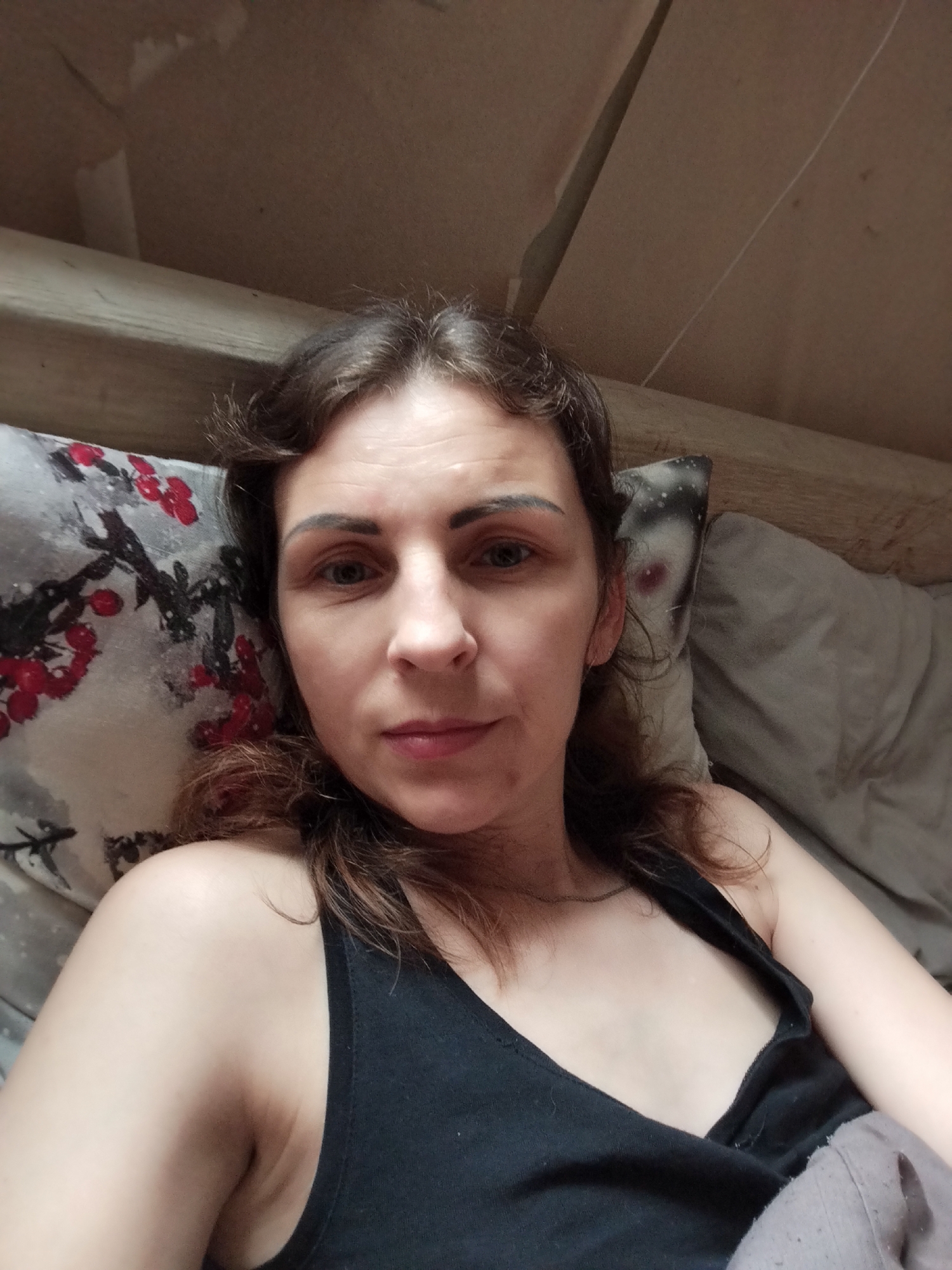 The image shows a medium shot of a woman lying on a bed, looking at the camera. She is wearing a black tank top. Her hair is brown and slightly wavy. She is lying on a pillow with a red and black floral pattern, and to her right is a gray pillow. The background appears to be a wooden structure, possibly a bed frame or ceiling. The lighting is dim. The image was taken in Vinnytsia, Ukraine.