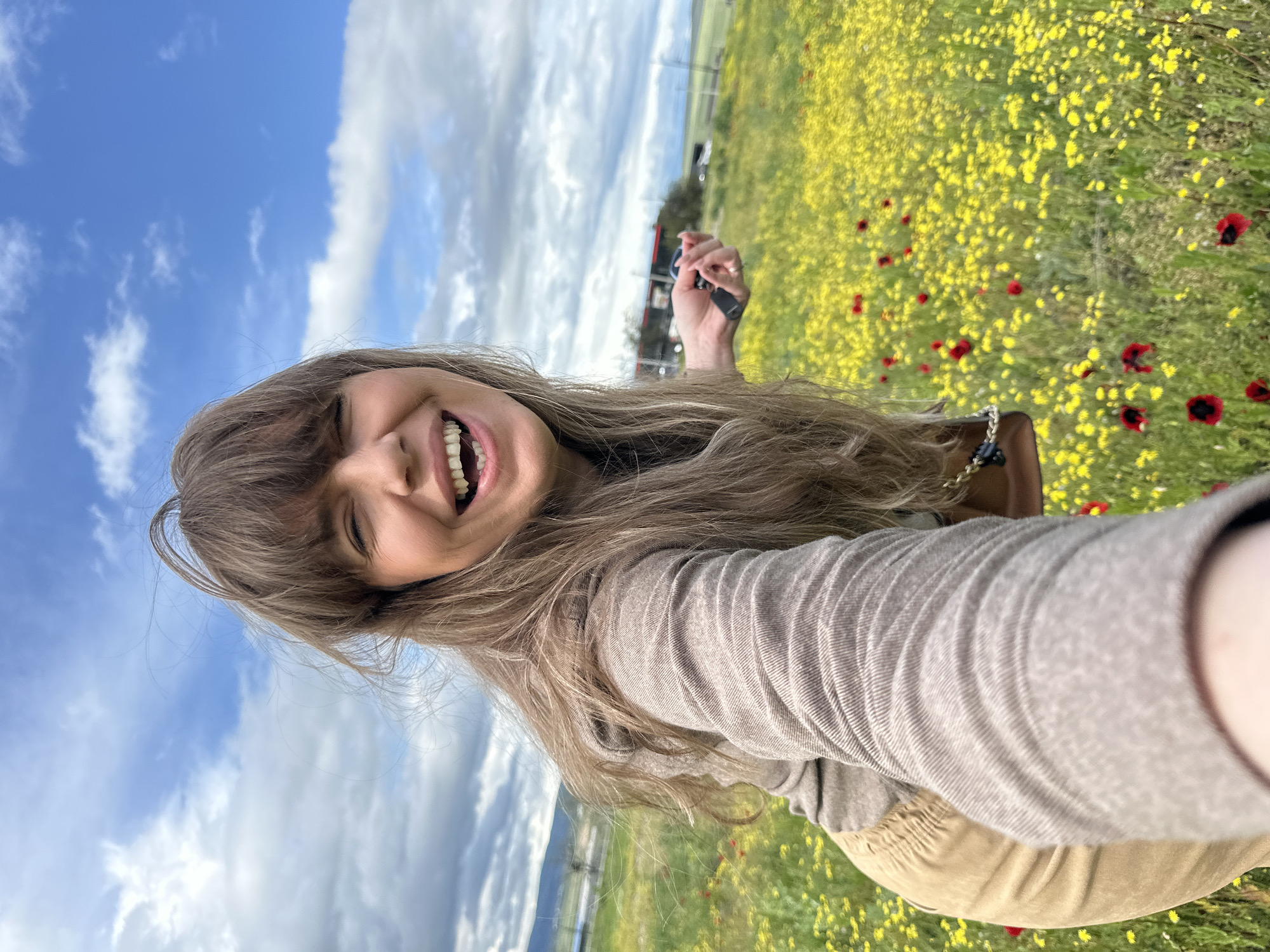 A woman with long, light brown hair and bangs smiles widely, revealing her teeth, in a selfie taken in a field of yellow and red flowers under a partly cloudy blue sky. She is wearing a long-sleeved beige shirt and khaki pants. Her right arm is outstretched, holding a small black object. A brown purse with a gold chain strap is visible on her right side. In the background, there are power lines, some trees, and a road with a vehicle. The location is likely in the Marneuli region of Georgia, based on the distinctive landscape of flowering fields and rolling hills.