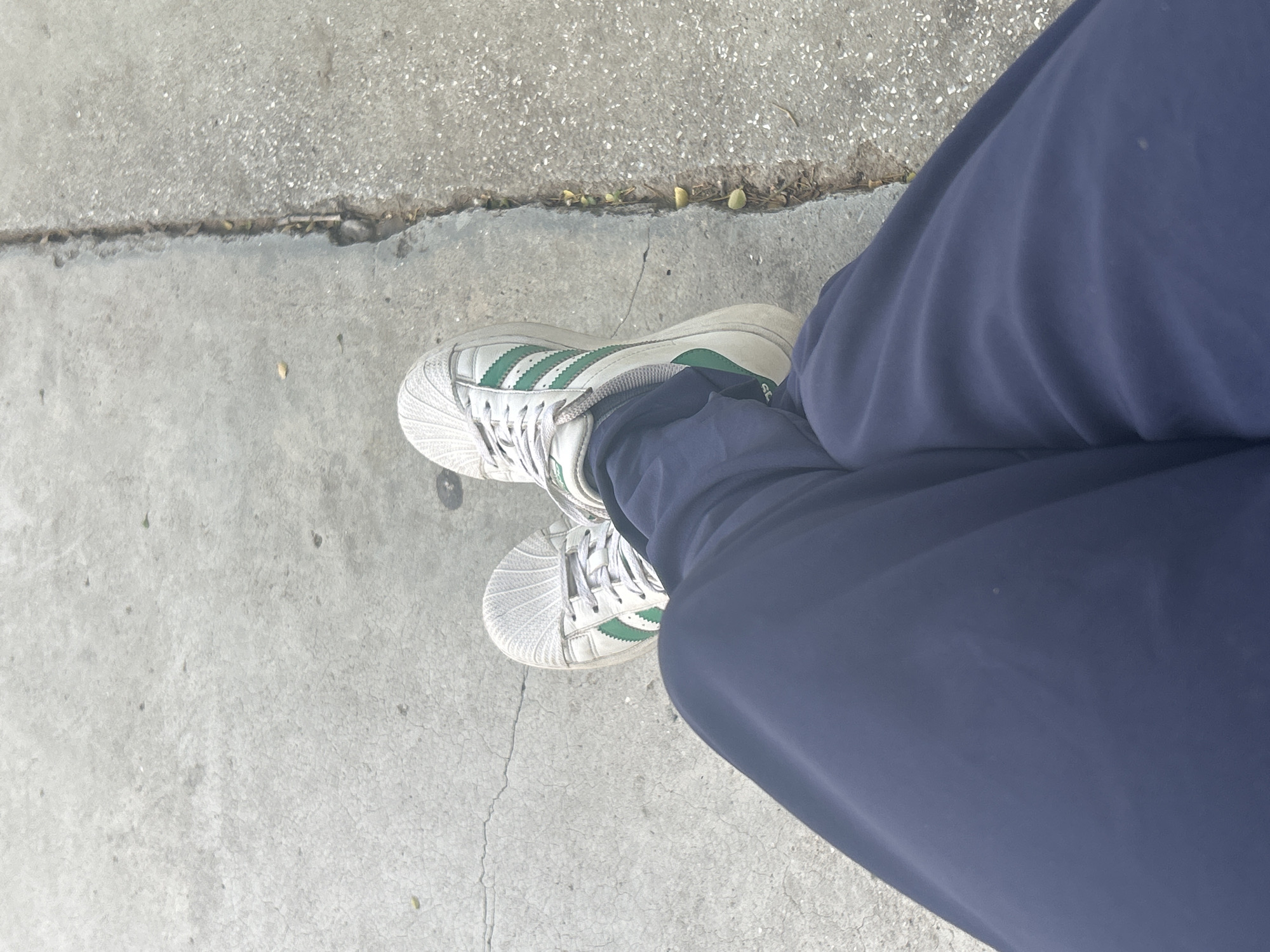 A person's legs, clad in dark blue pants and white Adidas sneakers with green stripes, are positioned with the right leg crossed over the left. They are resting on a cracked concrete surface, possibly a sidewalk or pathway, with a narrow gap running alongside. The environment appears to be outdoors, with the light suggesting it is daytime. The image does not provide enough information to determine the specific location within Santa Ana, Venezuela.