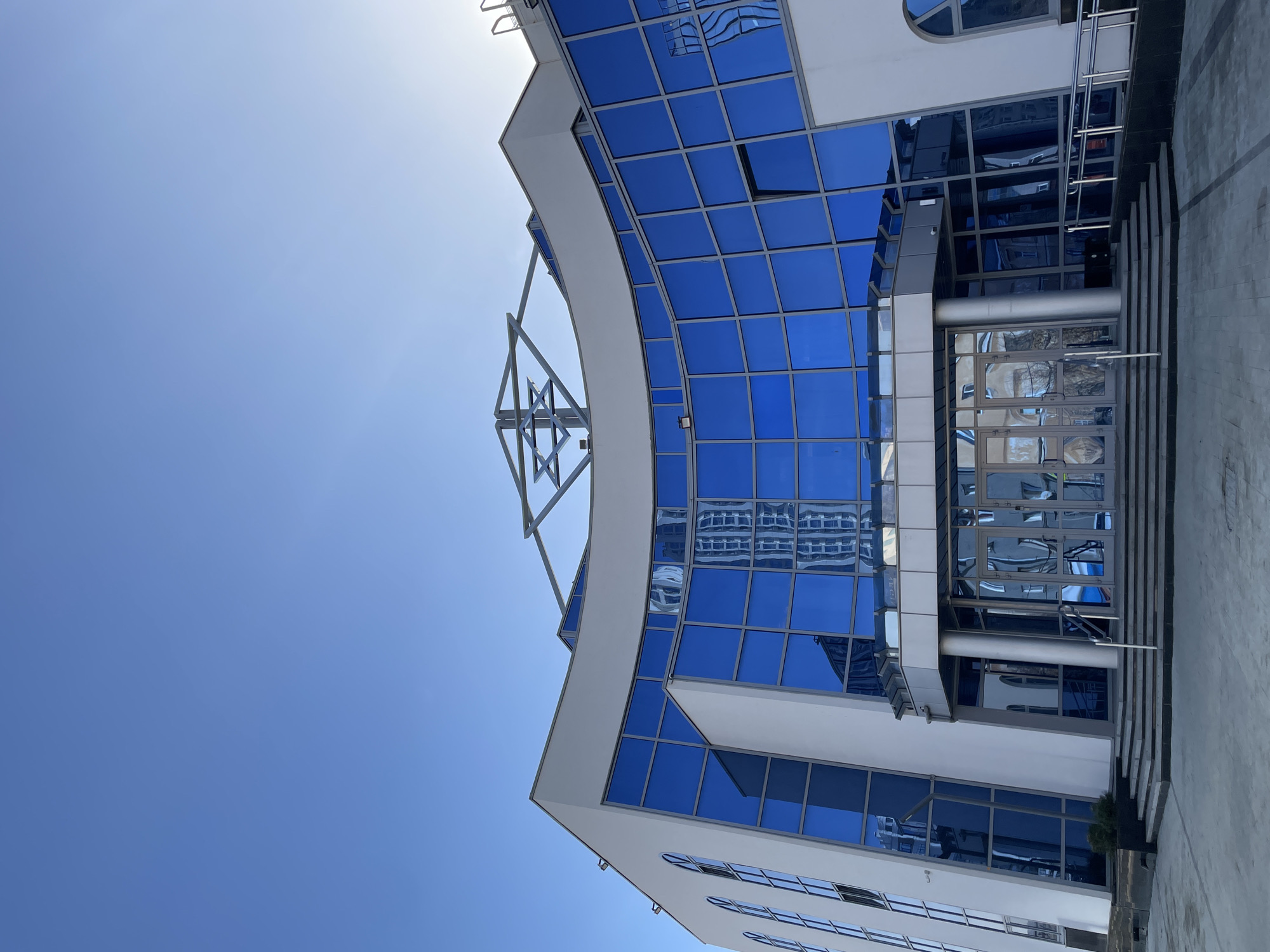 The image shows the exterior of a modern building in Yekaterinburg, Russia, with a curved façade of blue glass windows and white trim. The windows reflect other buildings. A metal structural support is partially visible above the curved section. The entrance features a set of glass doors and a staircase with metal railings. The sky is blue and the sun is visible in the upper left.