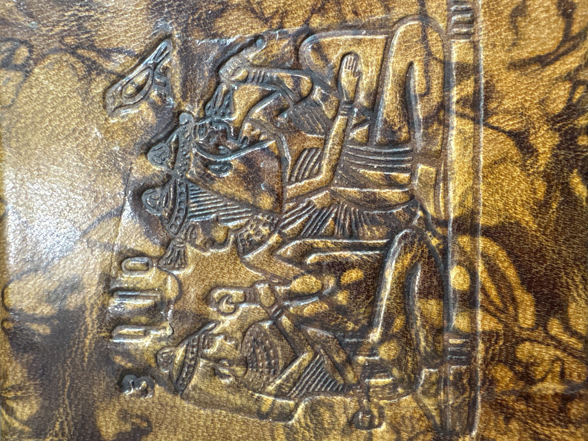 The image depicts a close-up of an embossed design on what appears to be leather. The design features figures in an ancient Egyptian style. One figure is seated and appears to be wearing a kilt, while another stands nearby with raised hands. Hieroglyphic symbols are also part of the design, next to the figures. The leather has a mottled brown and gold texture. The location in the city of Krasnoyarsk, Russia is not discernible from the image.