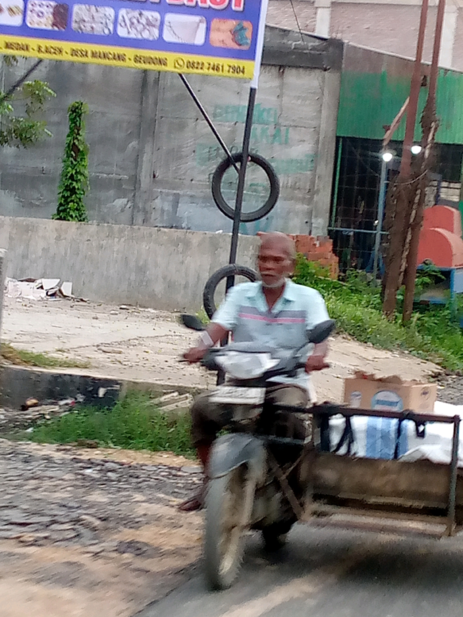 An elderly man with a shaved head and a goatee rides a motorcycle with a sidecar in Keudegeudong, Indonesia. The man wears a light blue polo shirt with pink stripes. He is driving on a paved road that appears to be in disrepair, with gravel and debris scattered around. The sidecar is laden with packages wrapped in brown paper and white cloth. In the background, a concrete wall and some overgrown vegetation are visible. A banner hangs above the wall, displaying text in Indonesian and a phone number. The overall scene suggests a busy, perhaps rural, commercial area.