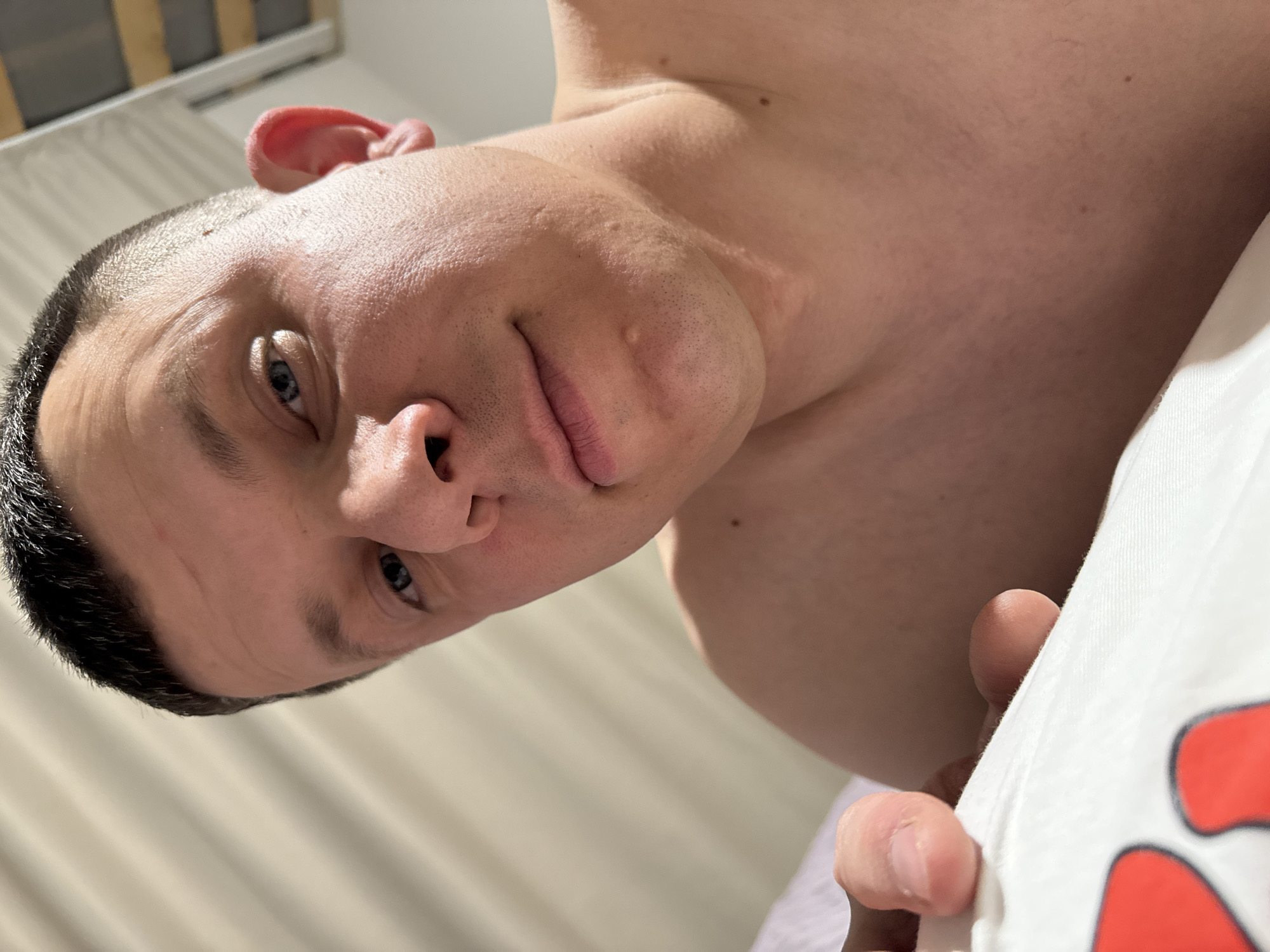 A close-up, slightly tilted shot of a man's face and upper chest. He has short, dark hair and light skin with visible pores and some minor skin imperfections. His eyes are blue. He is looking directly at the camera with a slight smirk. He is wearing a white t-shirt with red designs that is partially visible. The background includes a light-colored wall and a white curtain. The location is Gornyi, Russia.
