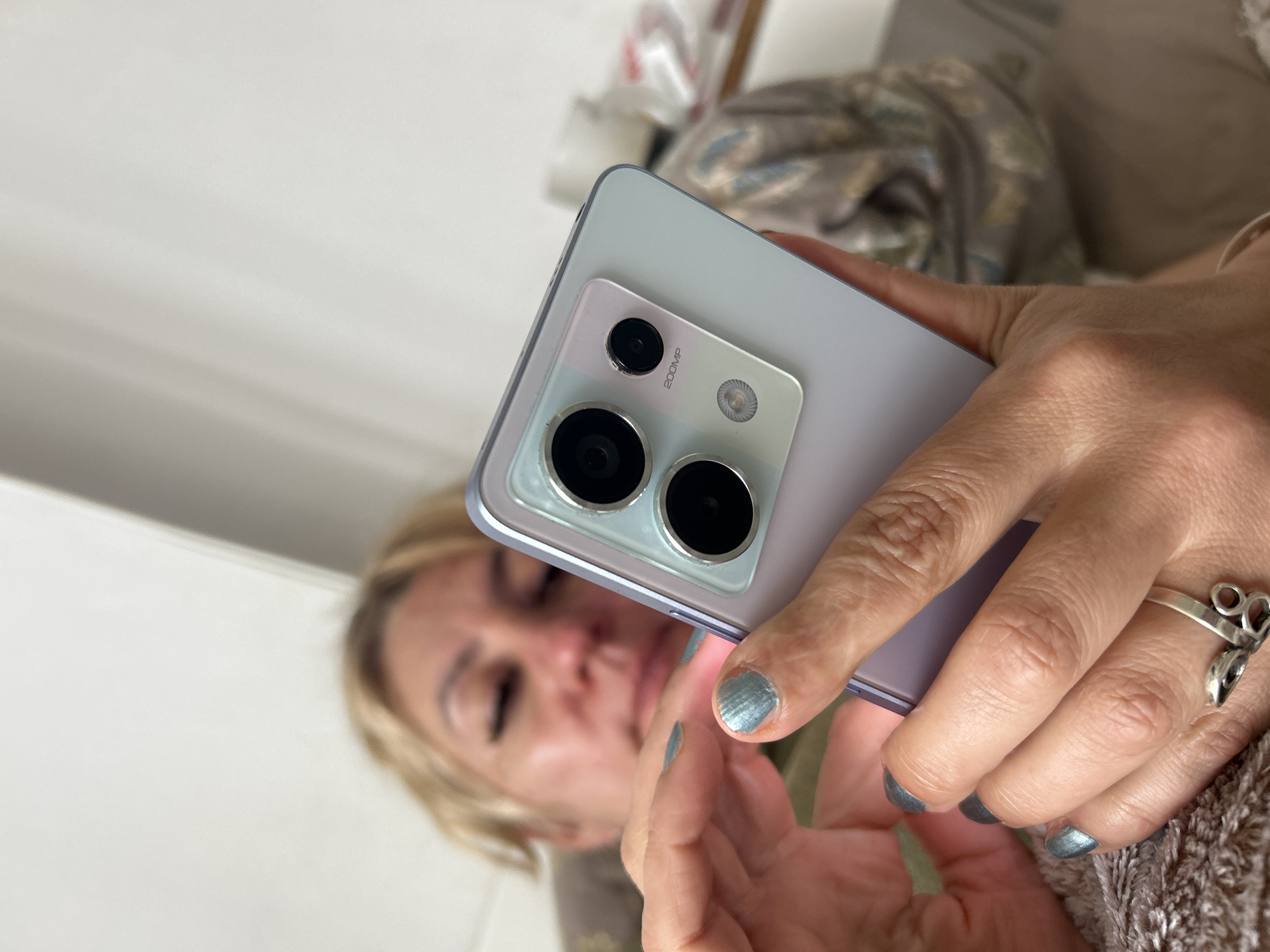 A woman is holding a smartphone in her hands. The smartphone has a distinctive camera array on the back, featuring a 200MP designation. The woman is out of focus, and her blonde hair is visible in the background. She is wearing a silver ring on her finger. The background includes a blurred mix of white and beige surfaces, possibly part of a bed or furniture. There is no specific information to indicate a location in Sofia, Bulgaria.