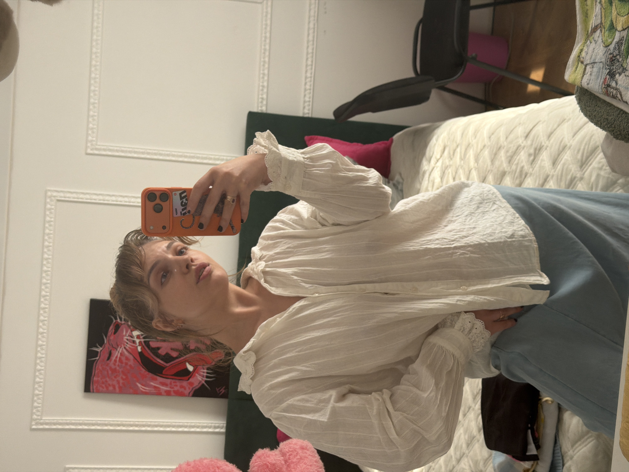A young woman takes a mirror selfie indoors in Большая Шильна, Russia. She is positioned centrally, holding an orange smartphone with a "PRADA" branded case in her right hand, capturing her reflection. Her gaze is directed towards the camera, with a neutral and calm expression.
She has light-colored hair, styled with bangs, and is wearing a loose-fitting, long-sleeved white blouse featuring textured fabric and delicate lace trim on the cuffs and collar. Light blue pants are visible below her waist. Rings adorn her fingers, and her nails are painted dark.
The background reveals a home interior, likely a bedroom or living space. White walls with decorative architectural molding frame the scene. A dark green upholstered headboard and a bed with a quilted white bedspread are prominent. To the right, a dark chair is visible, along with a pink cushion or yoga mat on the floor. A distinctive piece of artwork hangs on the wall behind the woman, depicting a stylized pink and red roaring animal face on a black canvas. Part of a patterned pillow or textile with a cartoonish design can be seen on the bed in the upper right corner. The lighting suggests it is daytime.