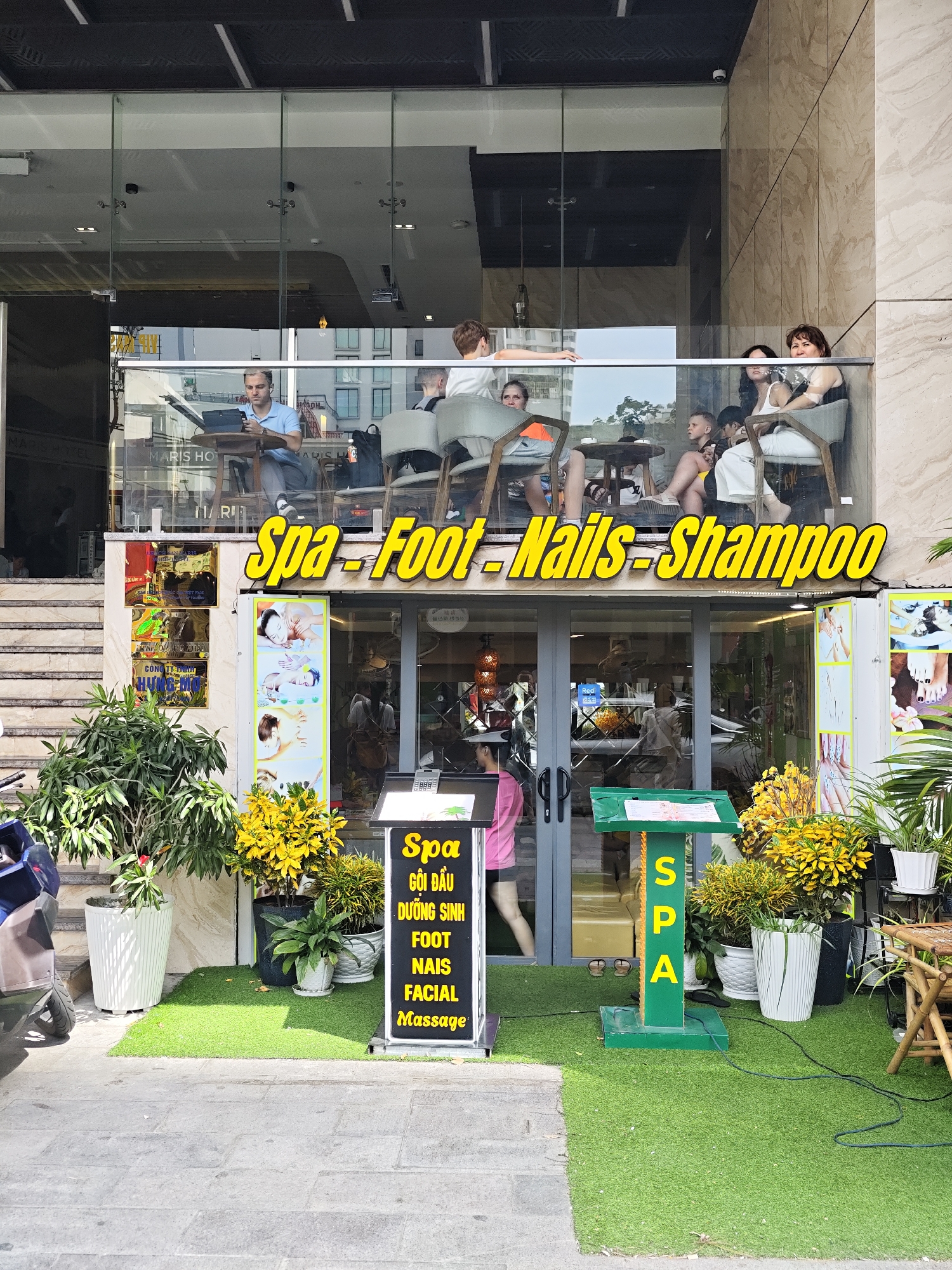 This image captures the exterior of a spa and salon located on a street in Nha Trang Ward, Vietnam. A prominent yellow sign with bold lettering reads "Spa - Foot - Nails - Shampoo." Below this sign, the entrance to the establishment is visible, with glass doors and windows reflecting the surroundings. To the left of the entrance, a set of stairs leads up to a higher level, where a balcony area is occupied by several individuals. One person is seated at a table with a laptop, while others are casually sitting and conversing.
On the ground level, in front of the spa entrance, a small patch of artificial turf is laid out. Several potted plants with lush green foliage and bright yellow flowers are strategically placed around this area. A green podium displaying the word "SPA" vertically in yellow letters stands on the turf. Another black sign with white text lists services such as "Spa," "Gội Dầu," "Dưỡng Sinh," "Foot," "Nails," "Facial," and "Massage." A motorbike is partially visible on the left side of the frame, suggesting it's a common mode of transportation in the area. The building facade features a modern design with large glass panels and light-colored stone cladding. The overall scene suggests a bright, sunny day.